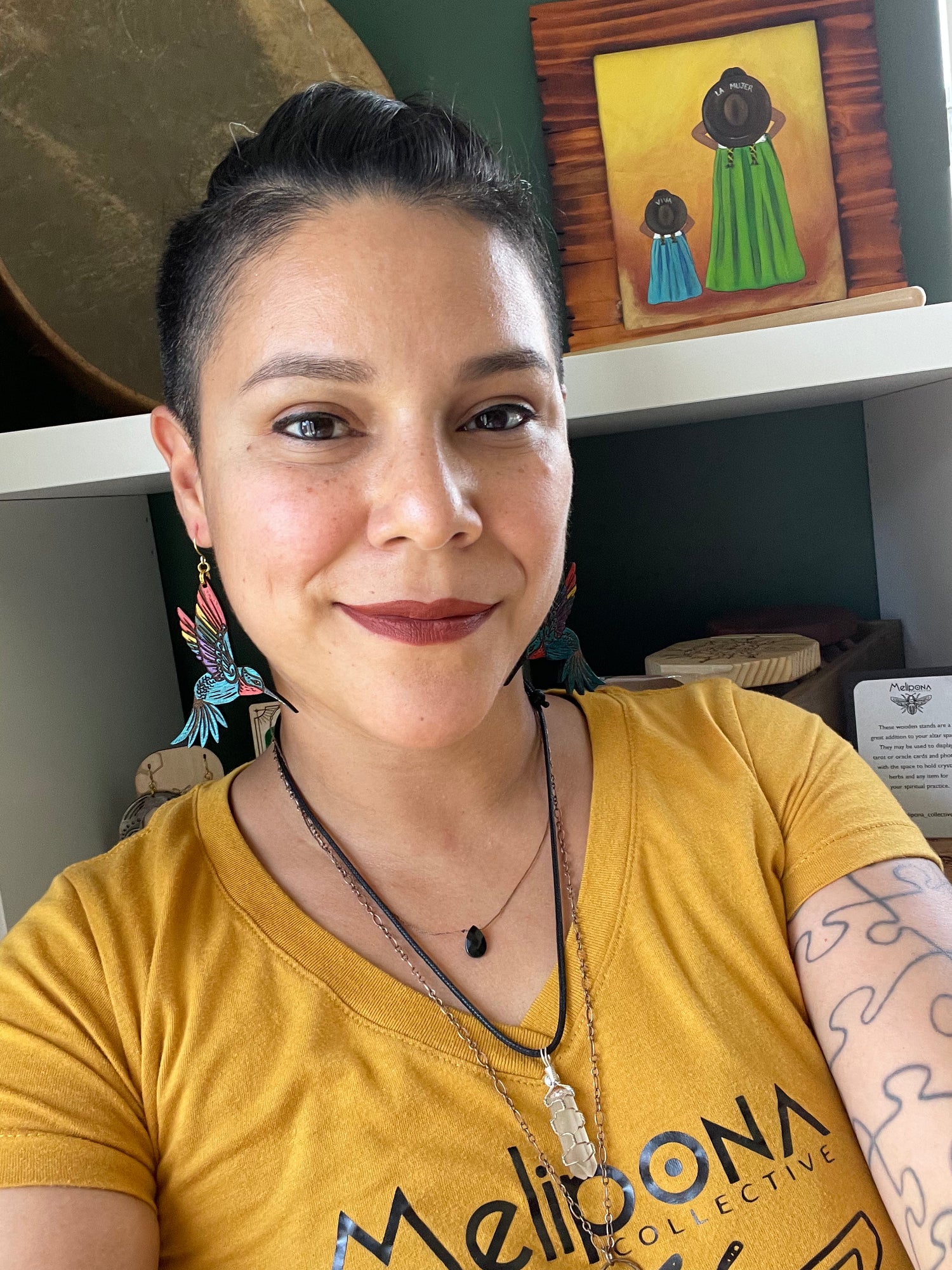 Crystal Castorena, queer mexican psychic medium, wearing a yellow shirt with the logo melipona collective and wearing blue hummingbird with rainbow wings earrings.