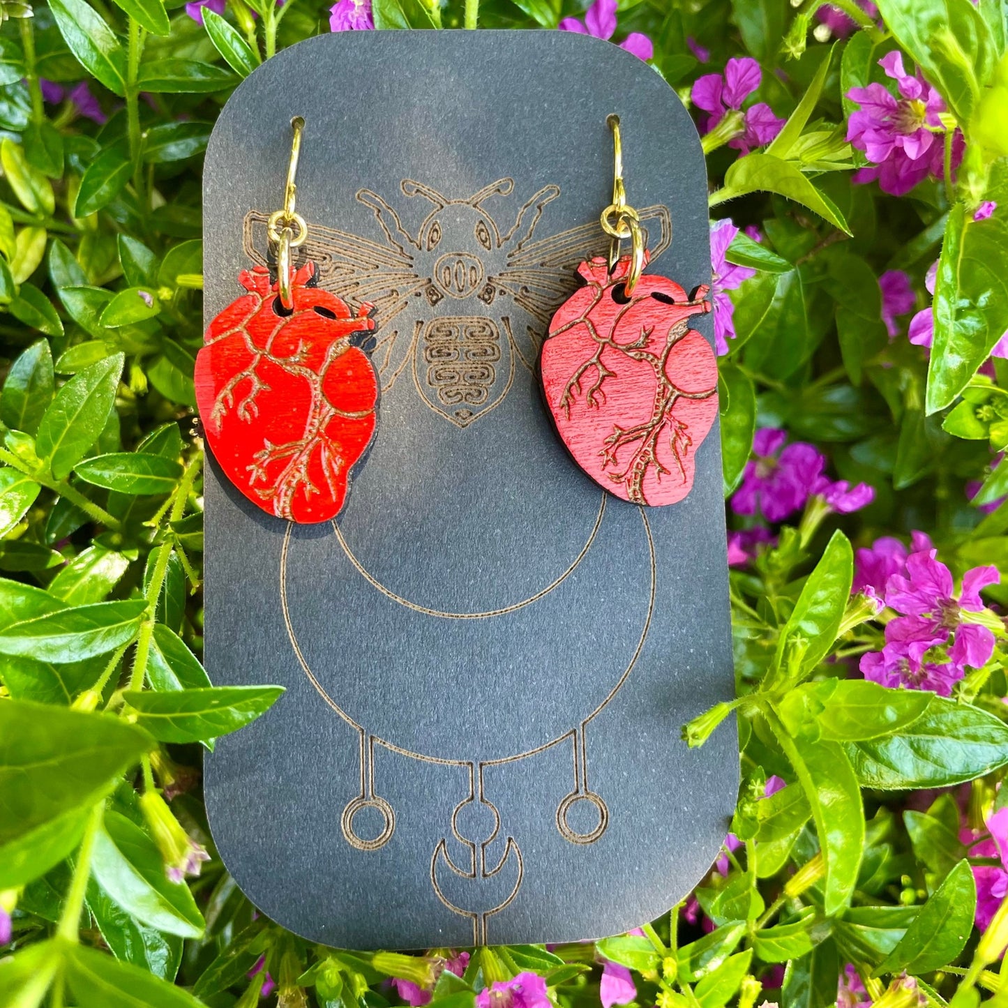 Red Wood Anatomical Hearts Earrings on gold colored hypoallergenic hooks on an earring card holder. Background green bush with purple flowers.