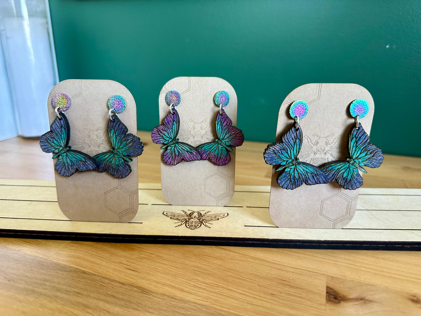 Three sets of Dark to light shades of Blue and purple Wood Butterfly Earrings on silver colored hoops and purple and blue glittery stud earrings.