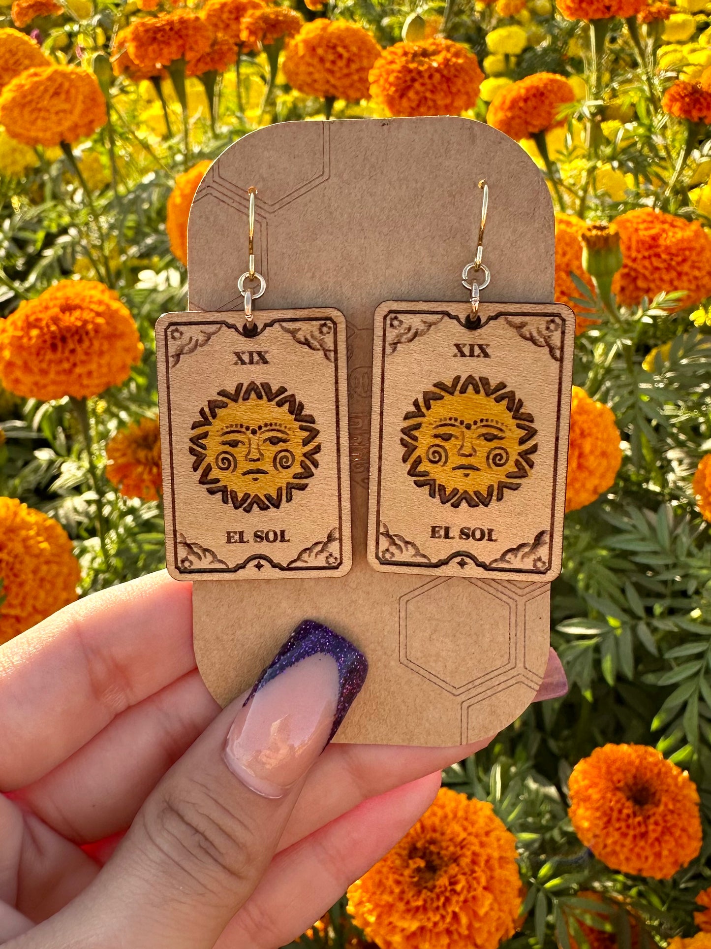 el sol the sun tarot card wood engraved earring with gold colored hypoallergenic hooks. Earrings are on a brown card stock earring holder being held in front of orange marigold field.