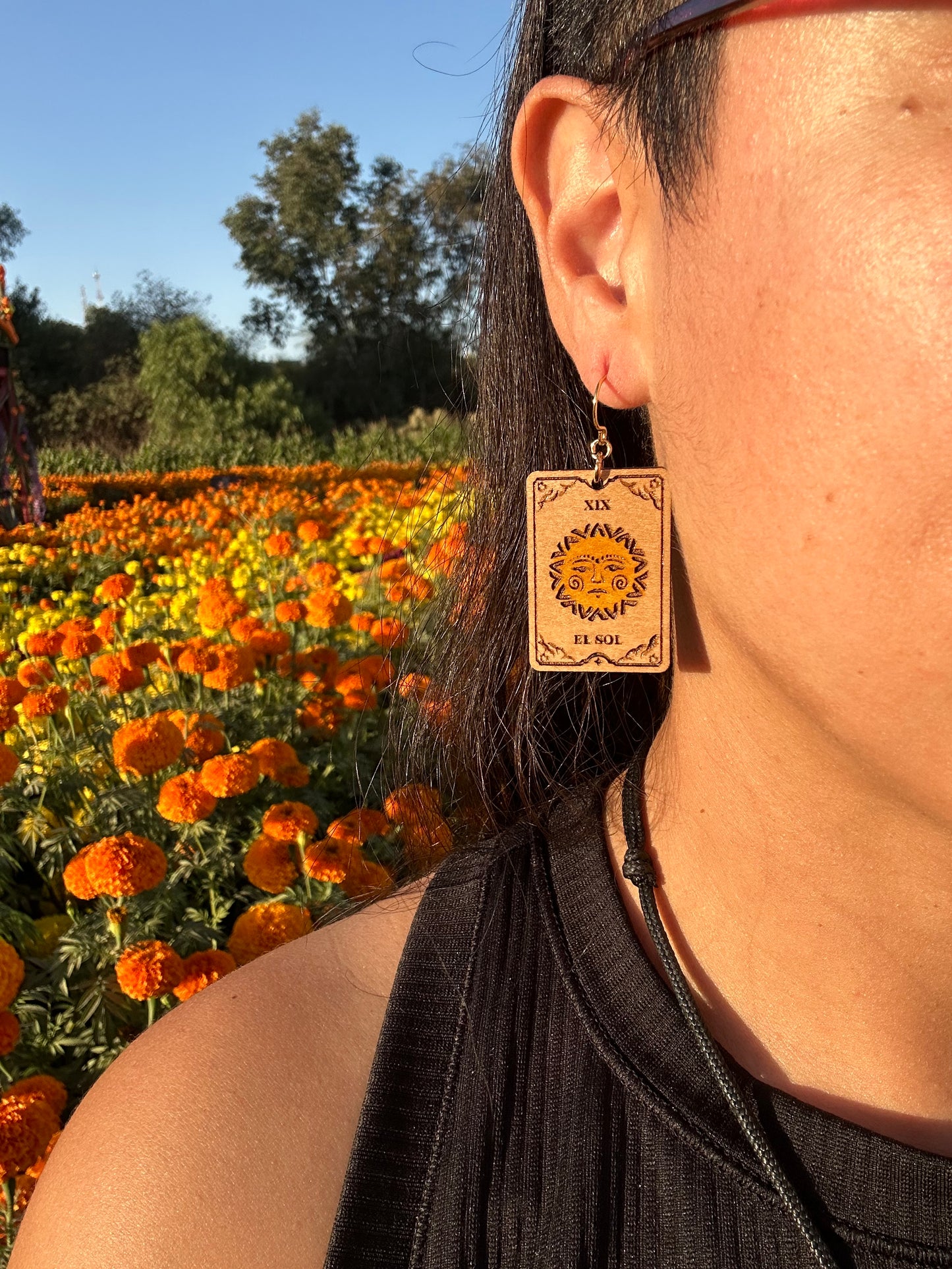 el sol the sun tarot card wood engraved earring with gold colored hypoallergenic hooks. Earrings are being worn by a woman close up visually only showing the side of her face and zoomed in to the ear wearing the earrings. Background is an orange marigold field.