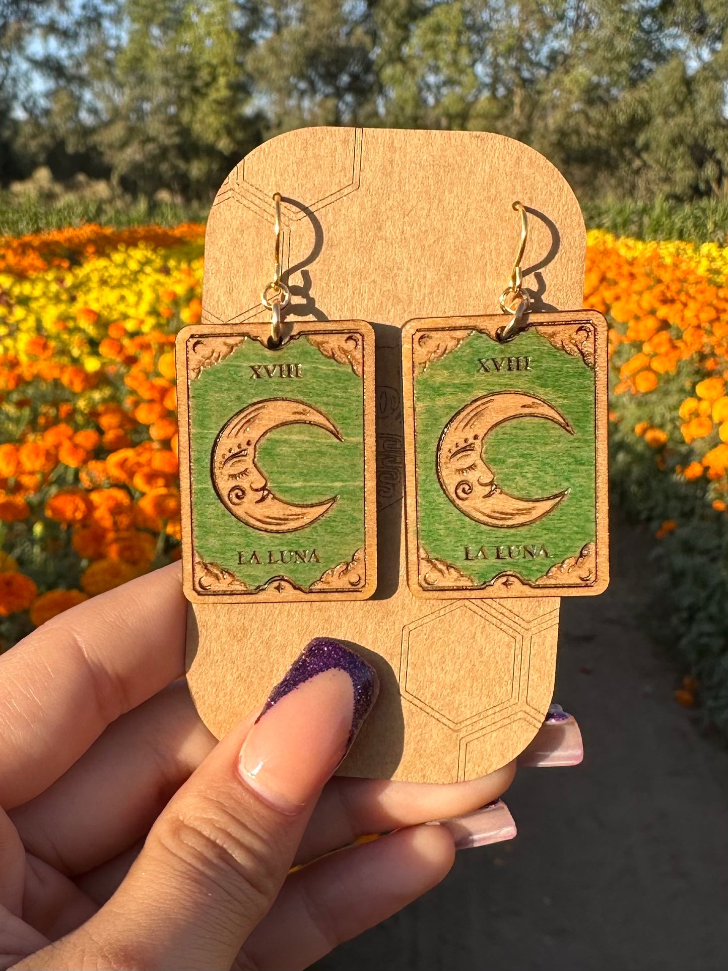 wood engraved la luna the moon tarot earrings on gold colored hypoallergenic hooks. Earrings on displayed on a brown card stock earring holder with a background of orange and yellow marigold fields.