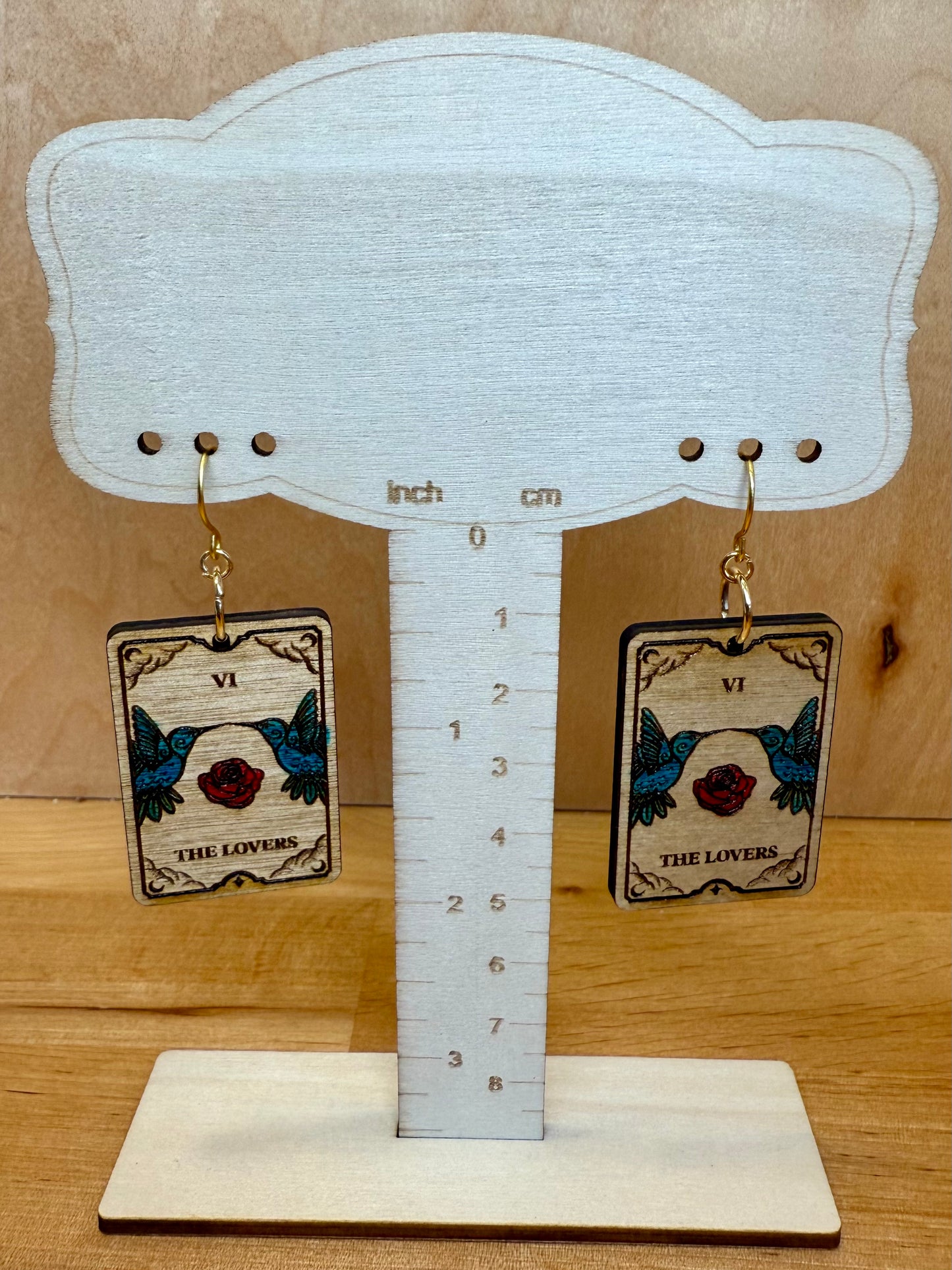 Earrings with 'The Lovers' tarot card design on a wooden stand with measurement scale.