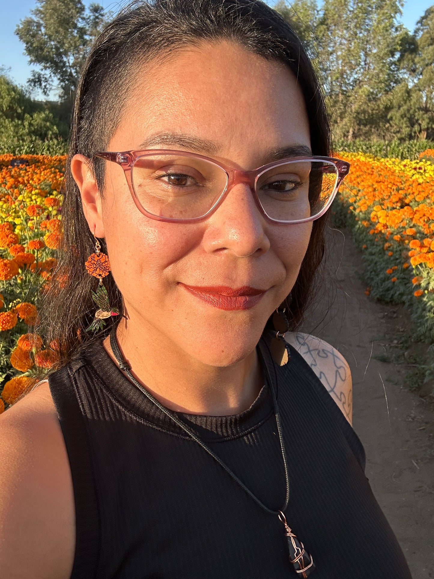 wood engraved earrings of an orange marigold with a green/white hummingbird with pink under beak. Earrings are on a gold colored hypoallergenic hook. Earrings are being worn by a women wearing glasses with a background of orange and yellow marigold fields.
