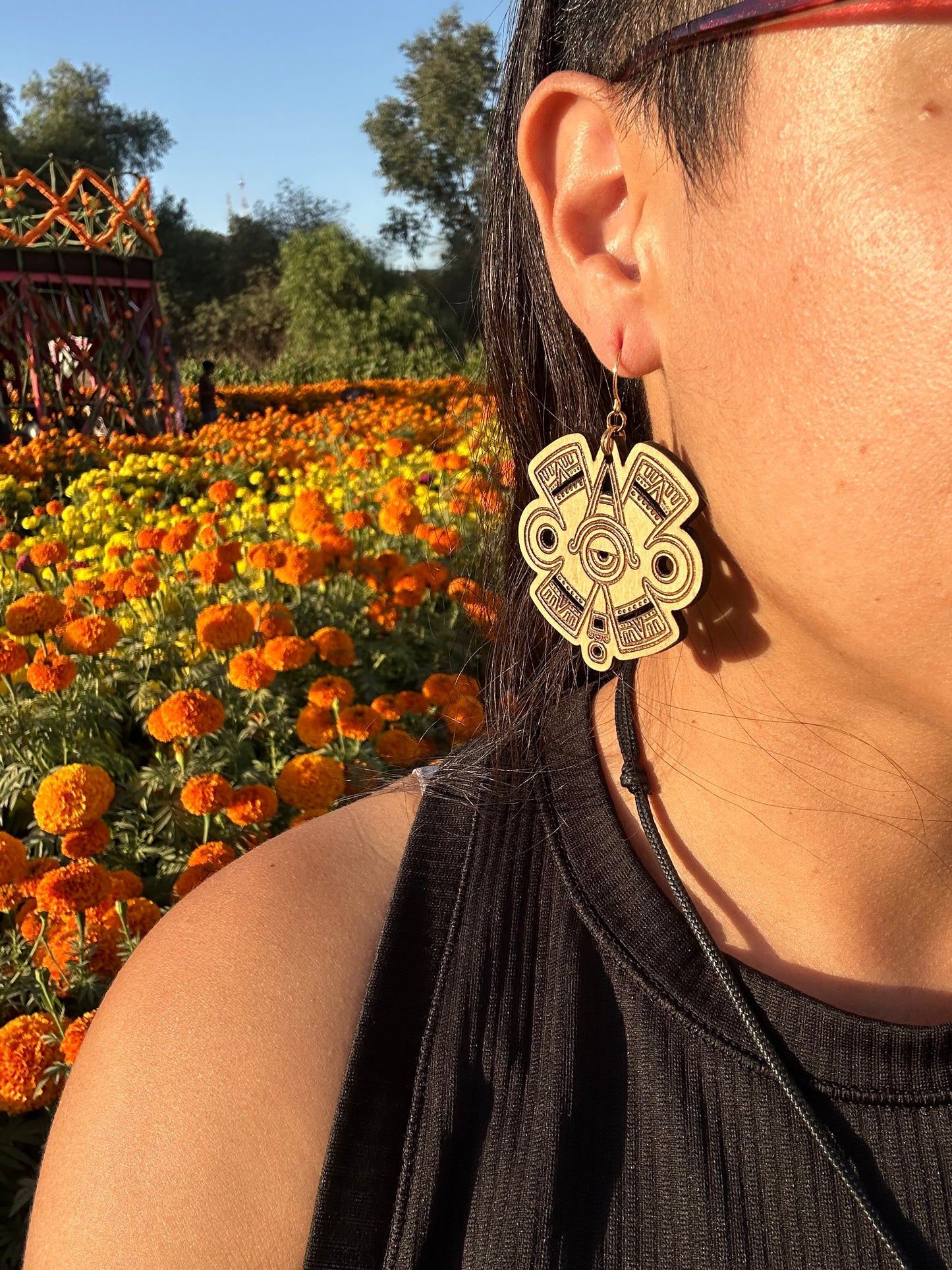 Ollin Earrings wood toned on gold colored hypoallergenic hooks. Earrings being worn by a woman wearing glasses looking off to the distance. Behind the woman is an orange and yellow marigold field.