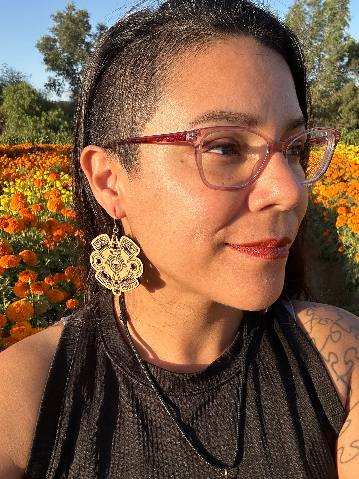 Ollin Earrings wood toned on gold colored hypoallergenic hooks. Earrings being worn by a woman wearing glasses looking off to the distance. Behind the woman is an orange and yellow marigold field.