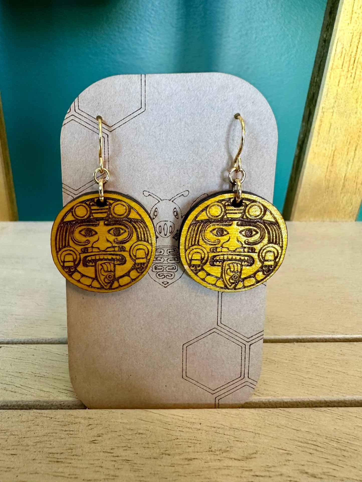 wood engraved Tonatiuh aztec sun yellow colored Earrings on a gold colored hypoallergenic earring hook. earrings on a brown card stock earring holder.