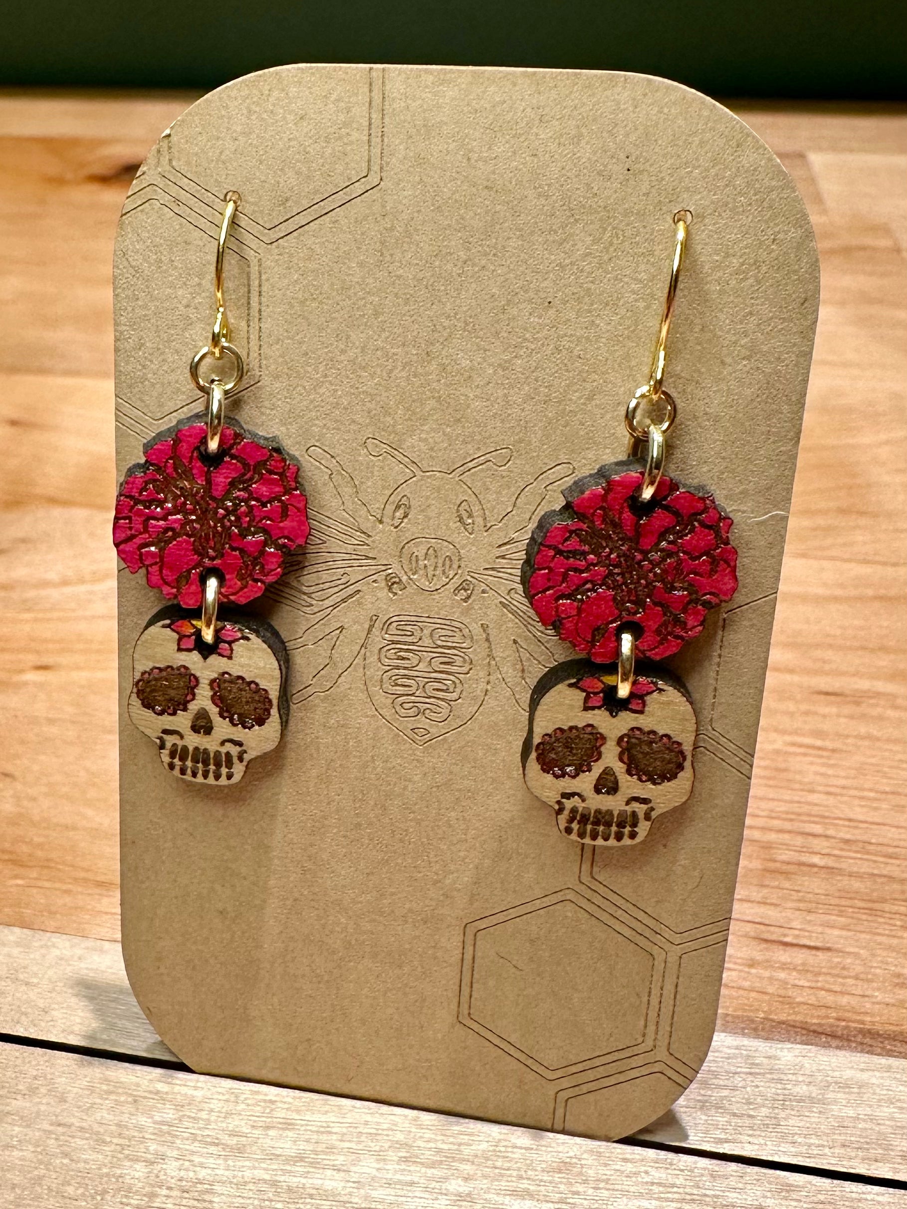 wood engraved pink colored rose with pink and wood tones sugar skull earrings on gold colored hypoallergenic earring hook. Earrings on a brown card stock earring holder. 