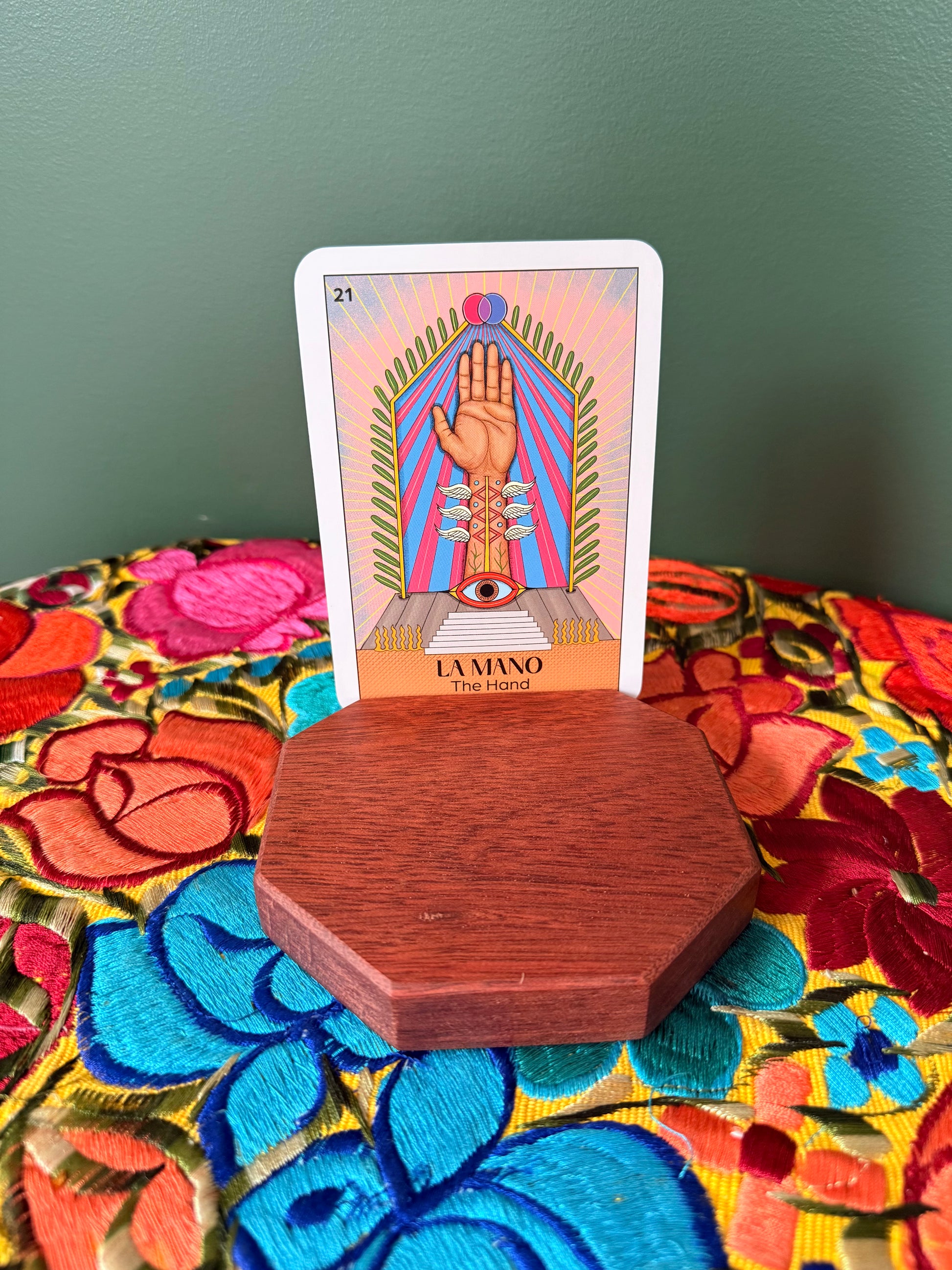 Tarot card titled 'La Mano' on a bloodwood wooden card holder altar with a colorful fabric background.