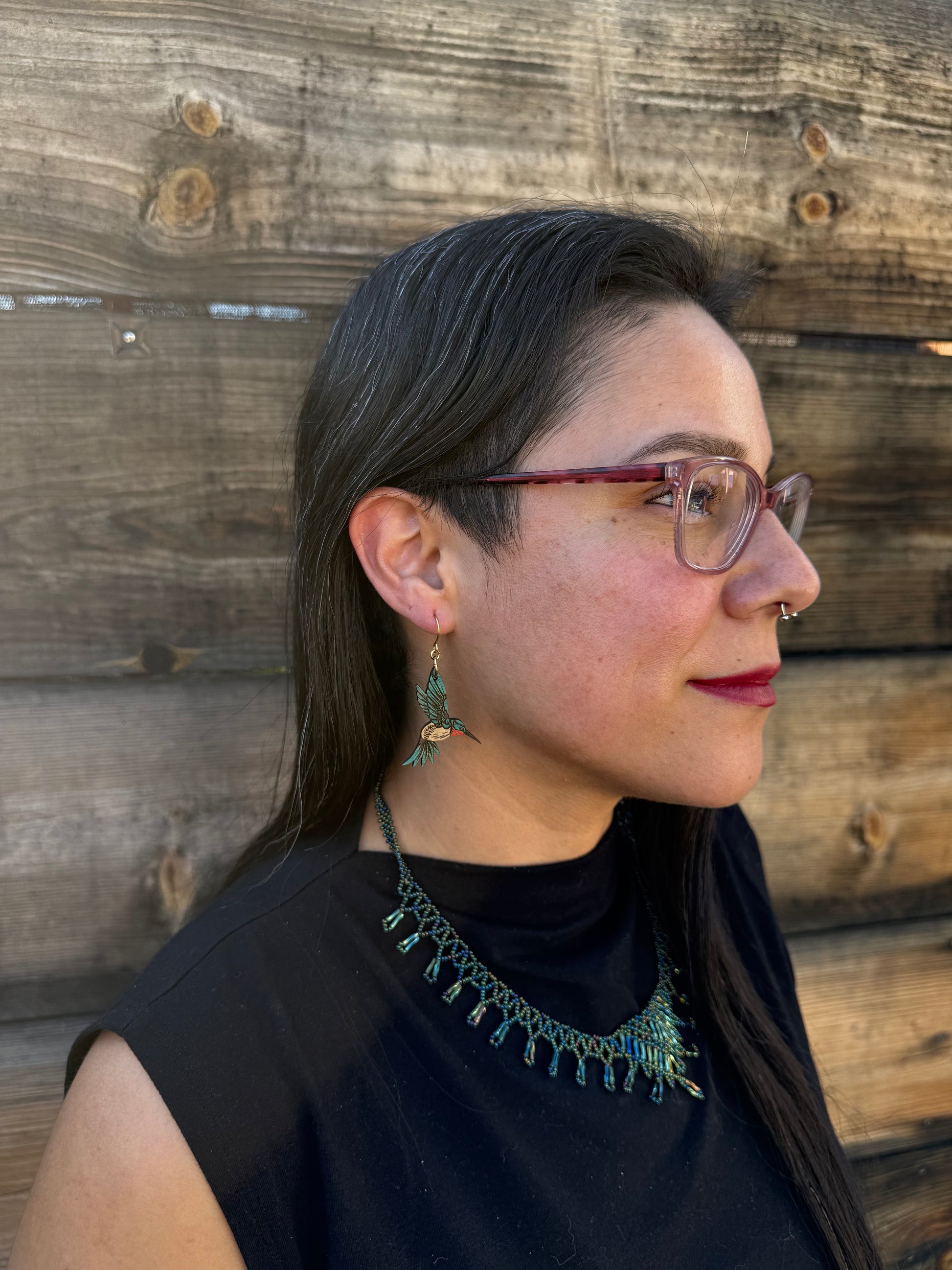 Woman wearing blue hummingbird earrings, glasses and a black top with decorative neckline on a wooden surface.