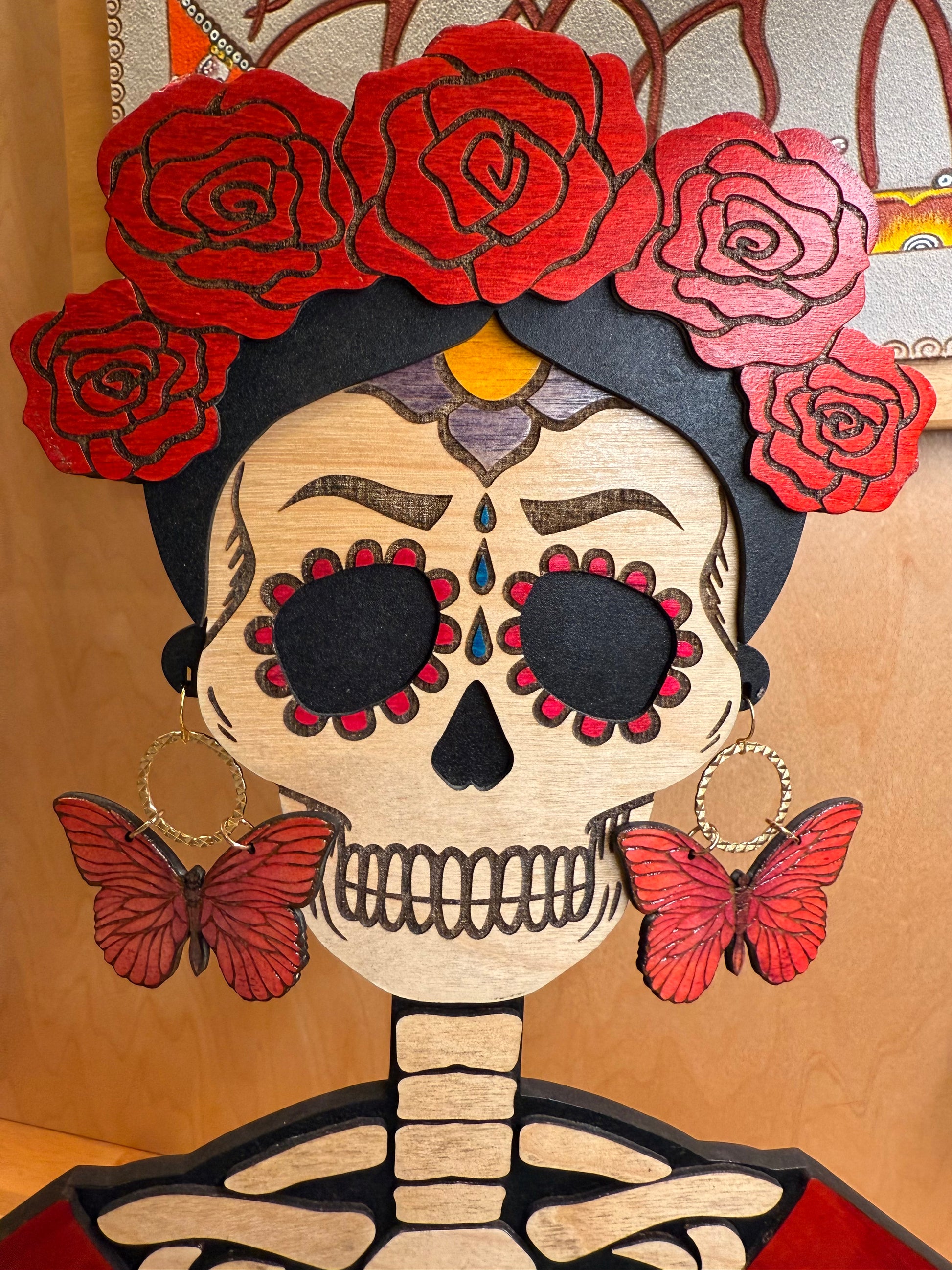 dark pink toned wood engraved butterflys dangling on a gold textured circle earrings being worn by a frida calevera wood statue.