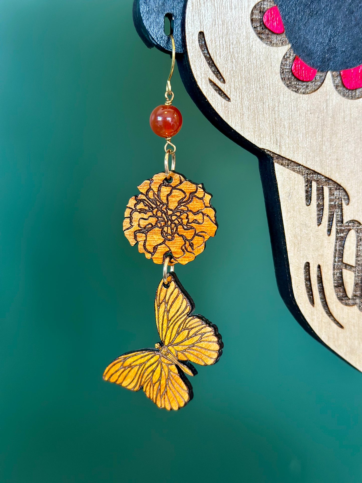 Orange marigold and yellow orange butterfly Wood earrings with orange carnelian crystal on gold colored hypoallergenic earring hooks. Earrings placed on a wood cut out design of a catrina skeleton woman ear hole close up. 