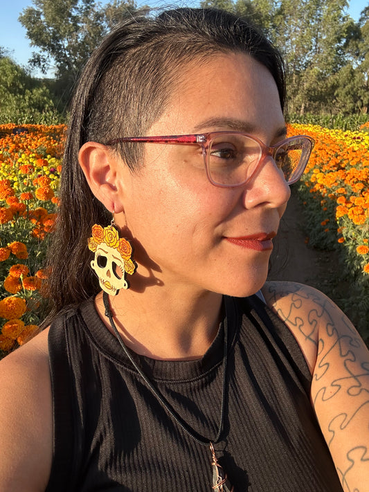 wood engraved catrina earrings with orange and yellow roses on head with gold coloredhypoallergenic earring hooks being worn by a woman wearing glasses looking to the distance. 