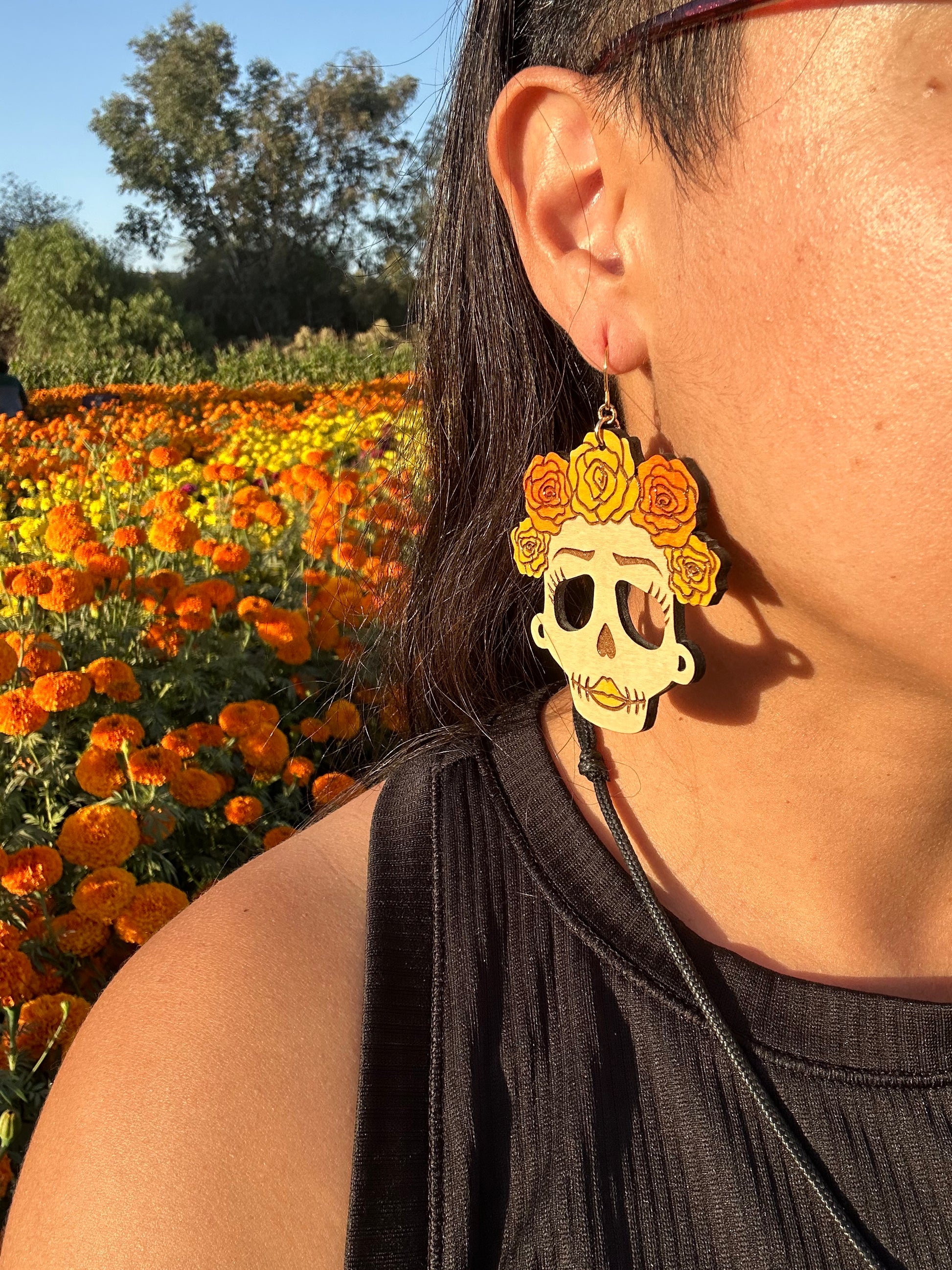 wood engraved catrina earrings with orange and yellow roses on head with gold colored hypoallergenic earring hooks being worn by a woman close up of ear with a background of a marigold field. 