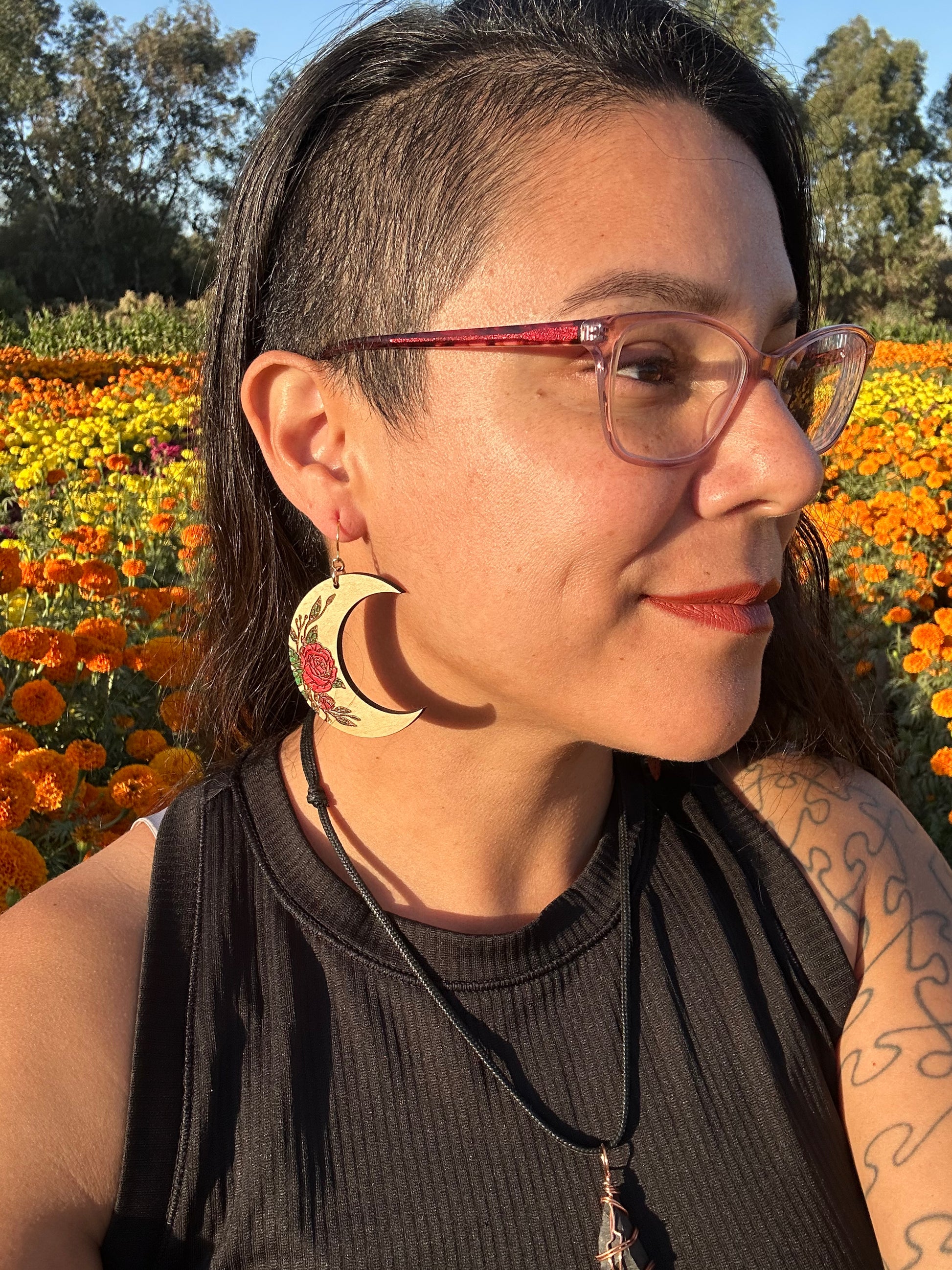 Wood engraved Crescent moon with red roses earrings on gold colored hypoallergenic hooks. Side profile of a woman with glasses wearing the earrings with a yellow and orange marigold field in the background.