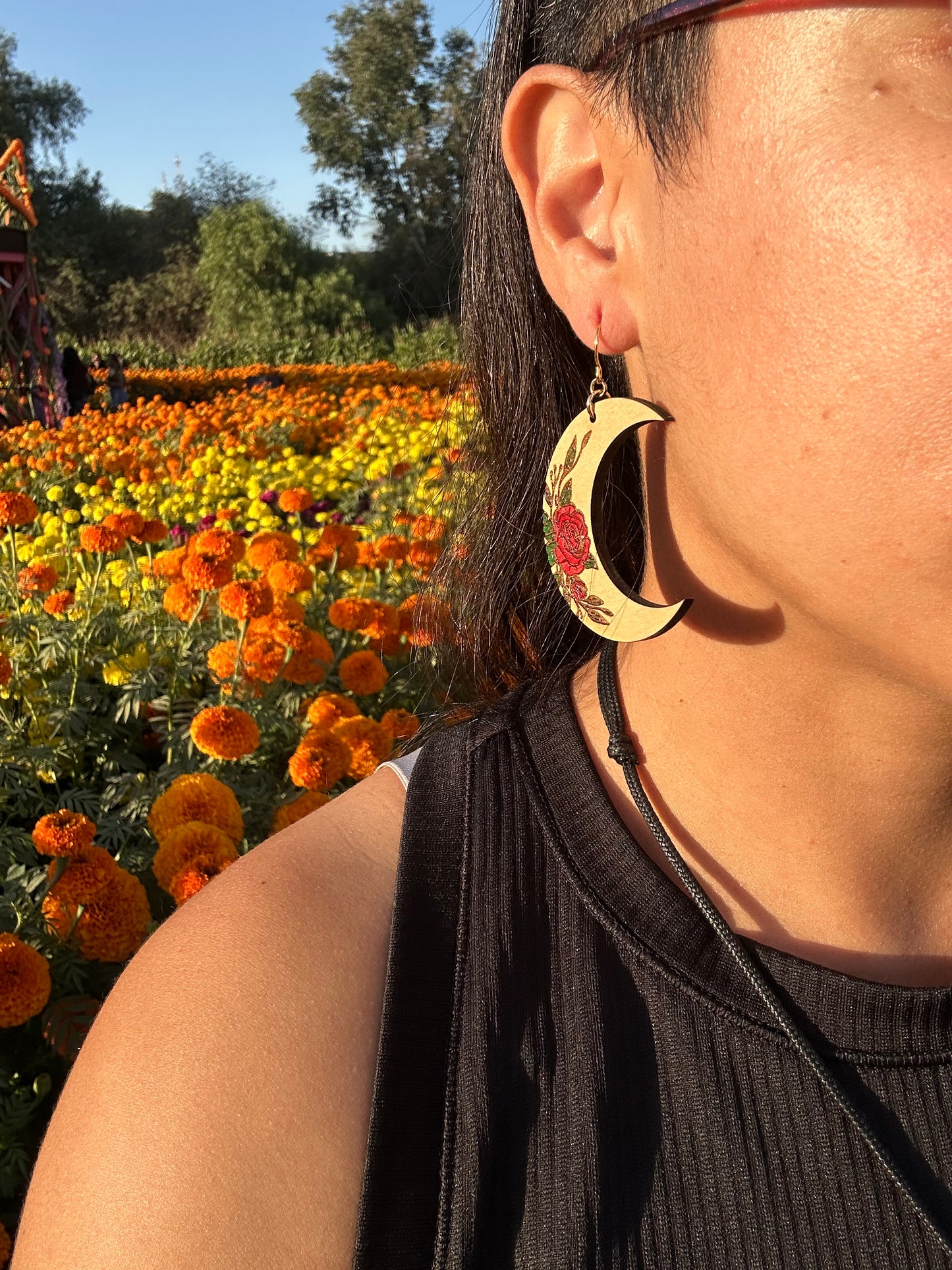 Wood engraved Crescent moon with red roses earrings on gold colored hypoallergenic hooks. Close up of ear of a woman wearing the earrings with a yellow and orange marigold field in the background.