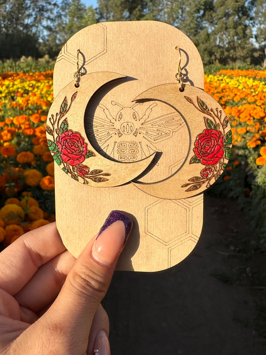 Wood engraved Crescent moon with red roses earrings on gold colored hypoallergenic hooks. Earrings on brown card stock earring holder being held with a yellow and orange marigold field in the background.