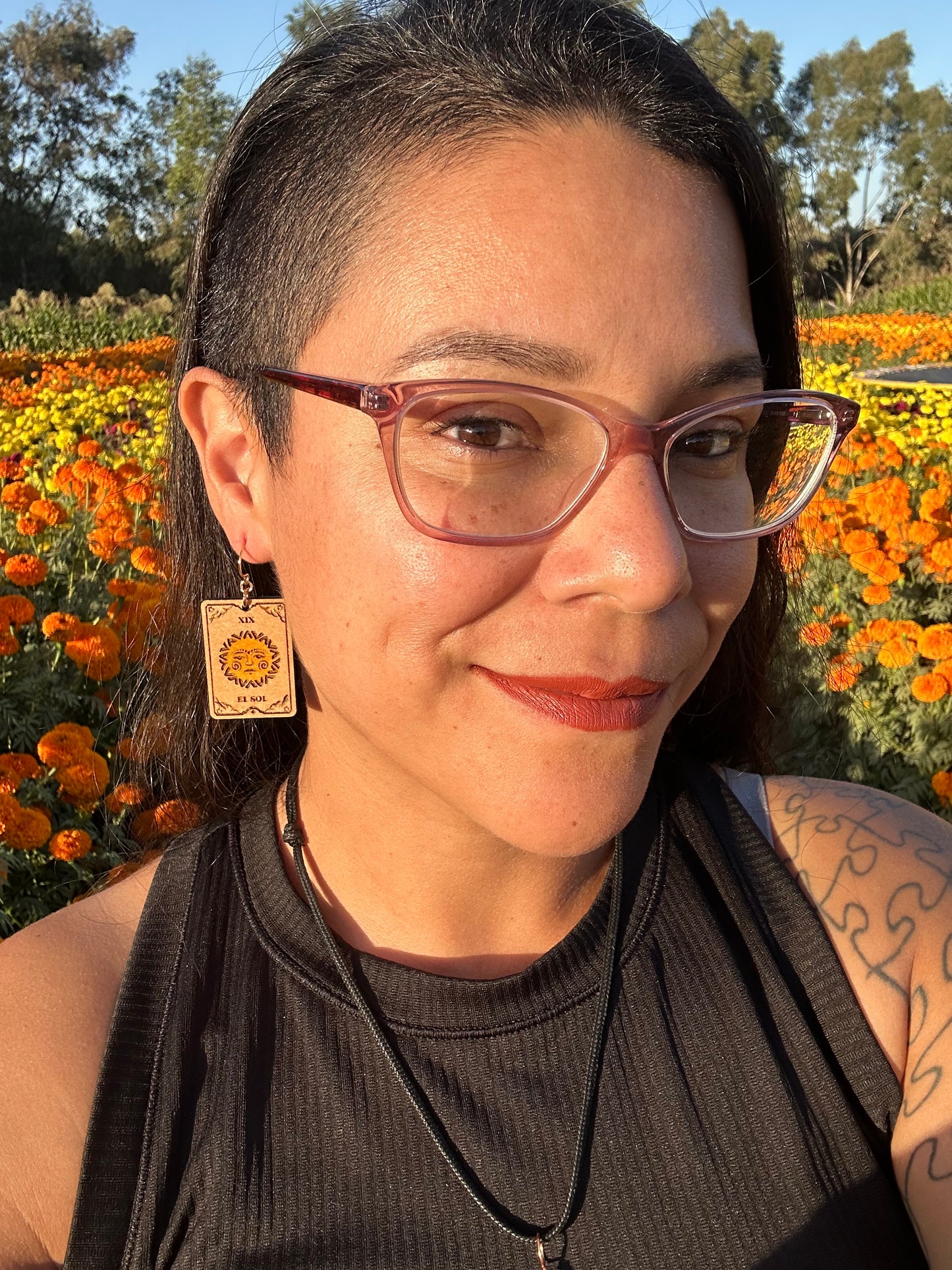 el sol the sun tarot card wood engraved earring with gold colored hypoallergenic hooks. Earrings are being worn by a woman wearing glasses in front of orange marigold field. 