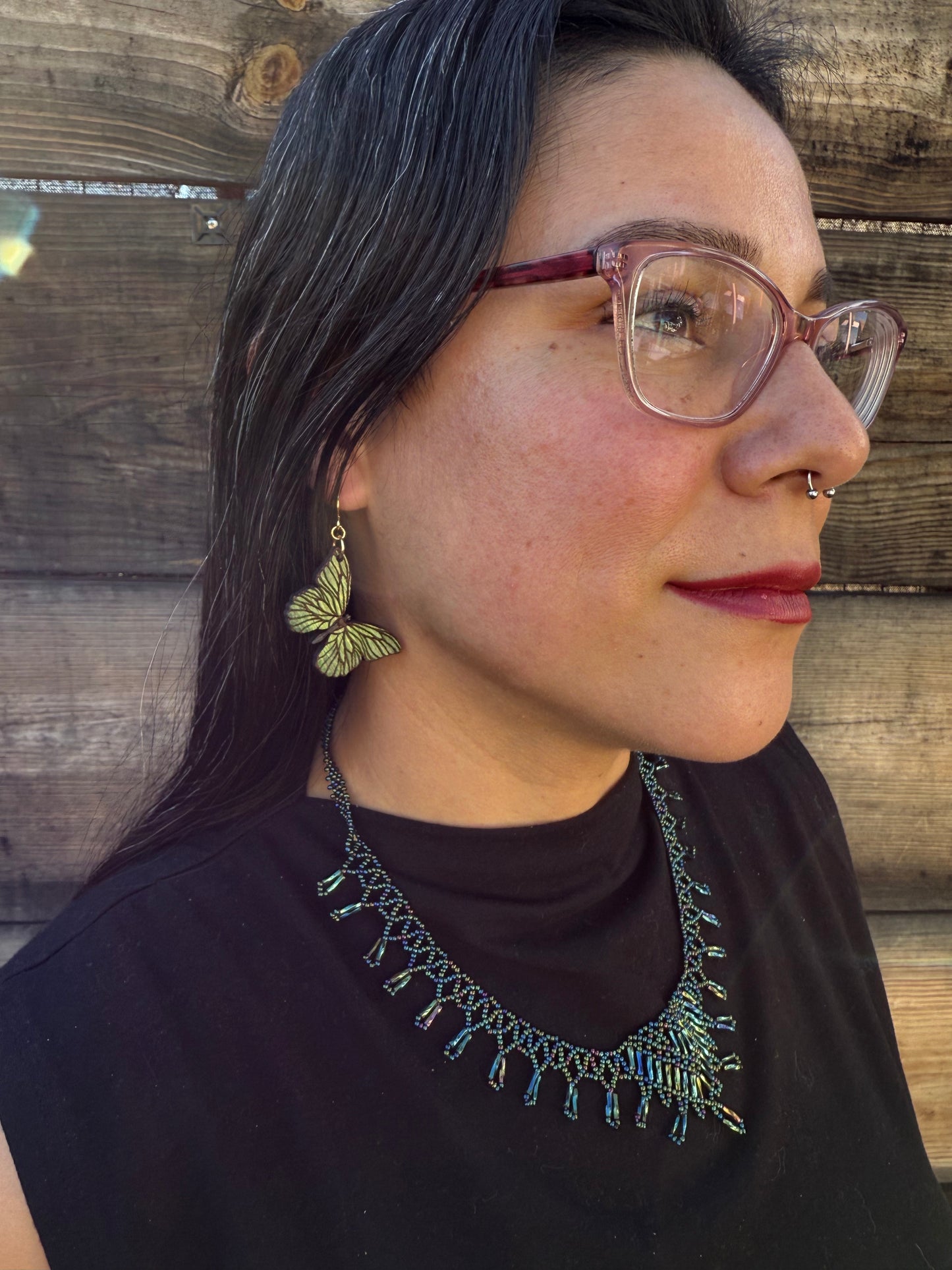 Person wearing glasses, green butterfly earrings and a dark top with decorative collar on a wooden surface.