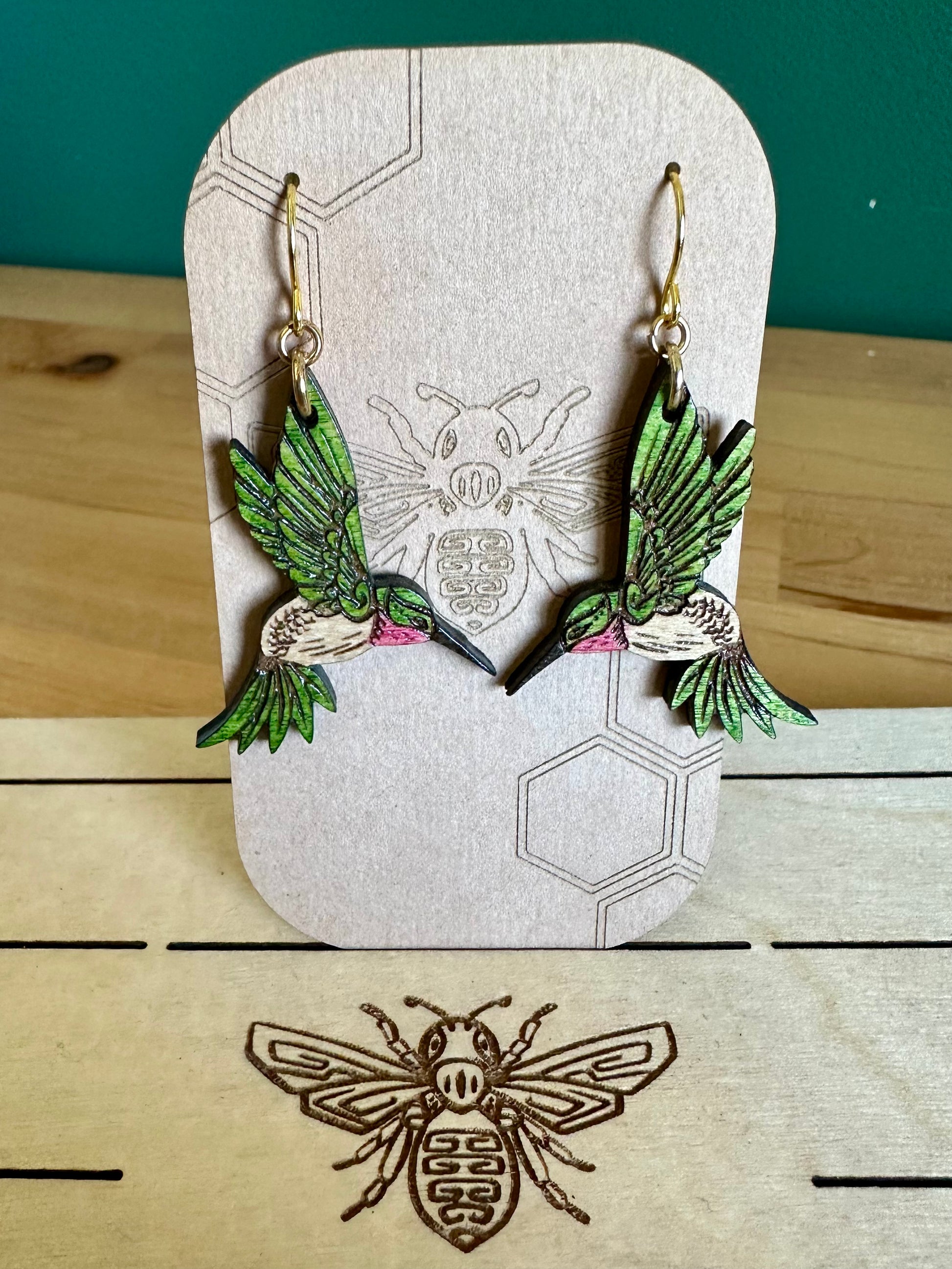 green, white hummingbird with pink under beak wood earrings using gold colored hypoallergenic hooks on a brown card stock earring holder. 