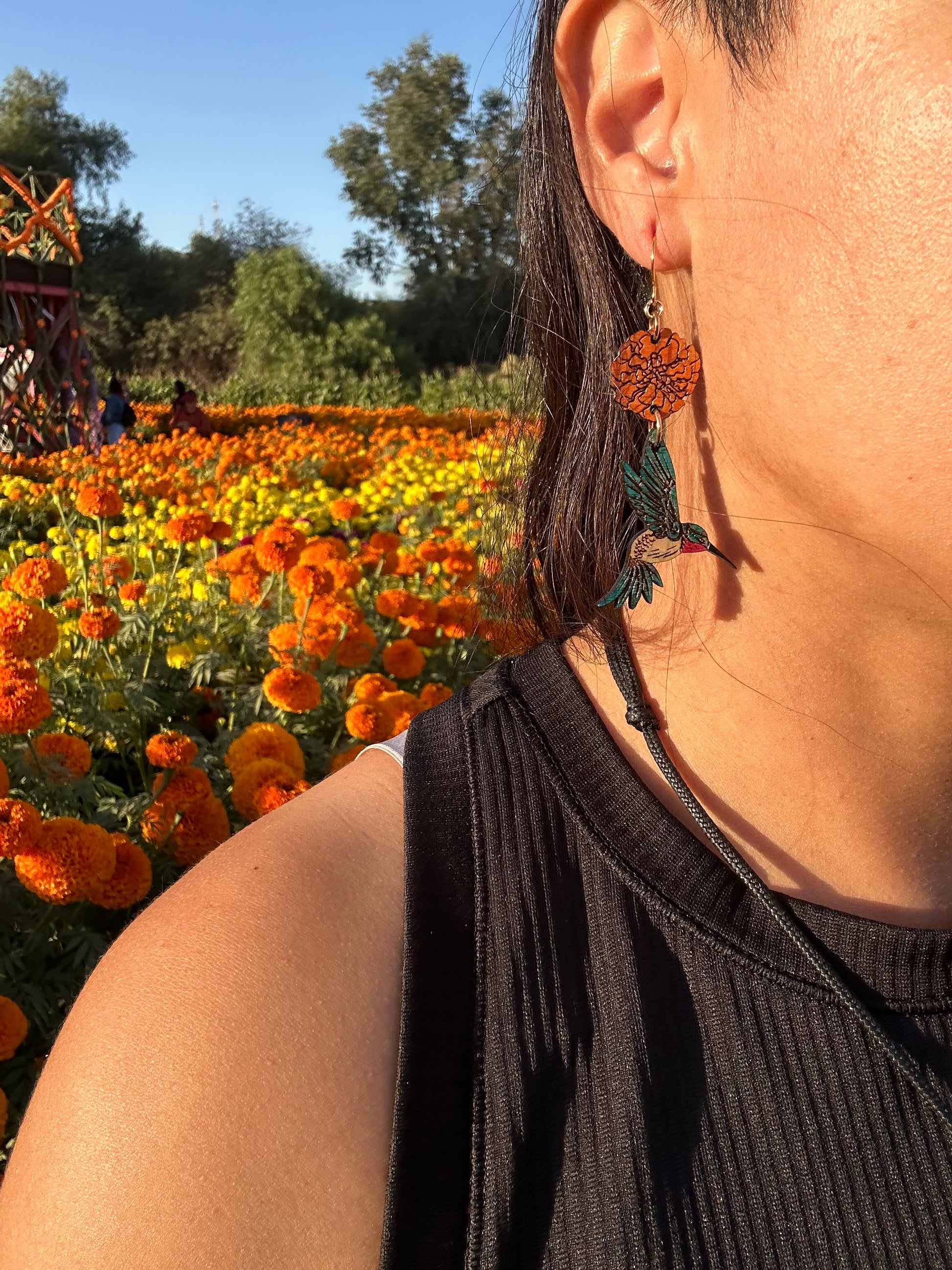 wood engraved earrings of an orange marigold with a blue/white hummingbird with pink under beak. Earrings are on a gold colored hypoallergenic hook. Earrings are being worn by a women with closeup of side profile and zoomed in on the ear with a background of orange and yellow marigold fields. 