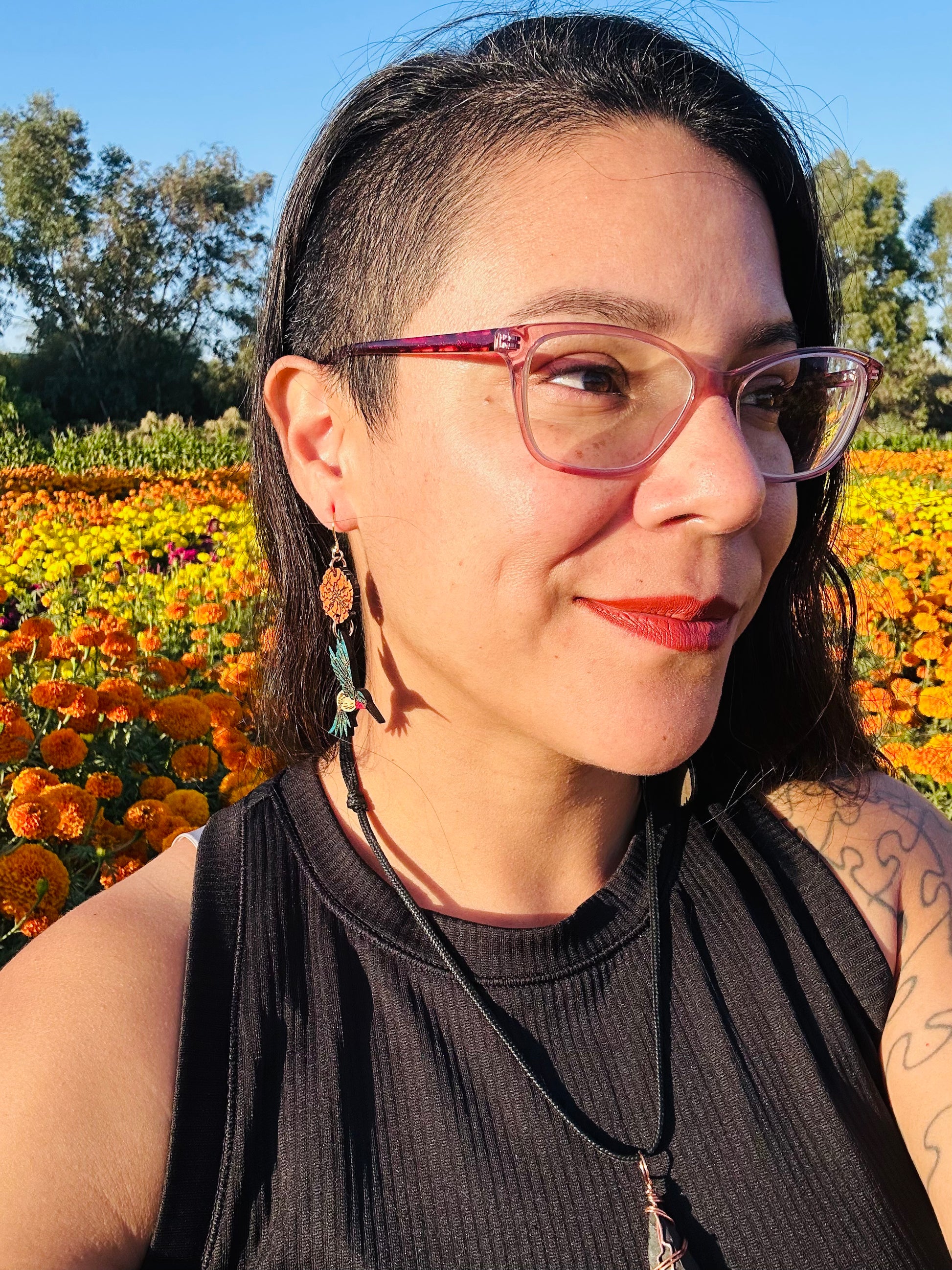 wood engraved earrings of an orange marigold with a blue/white hummingbird with pink under beak. Earrings are on a gold colored hypoallergenic hook. Earrings are being worn by a women with closeup of side profile with a background of orange and yellow marigold fields. 