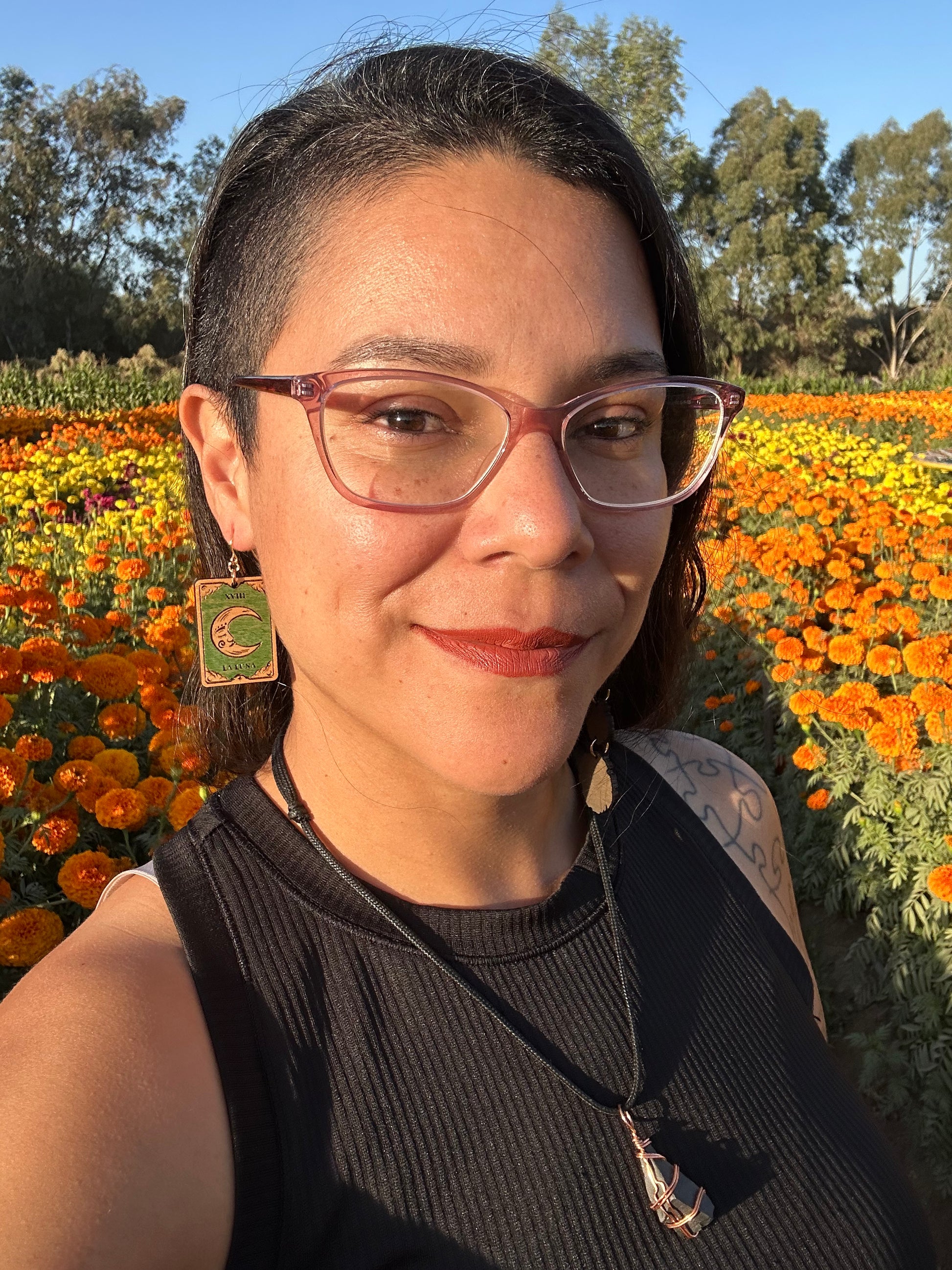 wood engraved la luna the moon tarot earrings on gold colored hypoallergenic hooks. Earrings being worn by a woman wearing glasses with a background of orange and yellow marigold fields. 