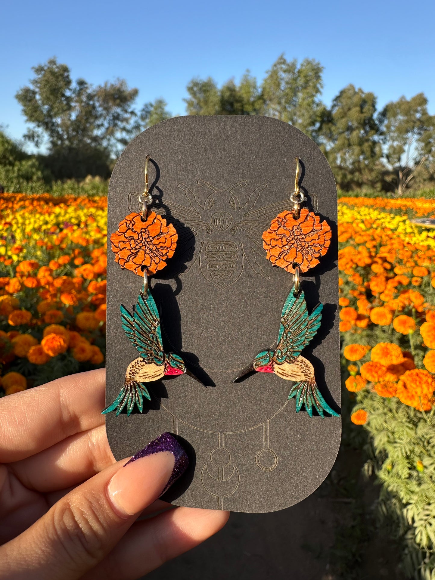 wood engraved earrings of an orange marigold with a blue/white hummingbird with pink under beak. Earrings are on a gold colored hypoallergenic hook. Earrings are being held on a black cardstock earring holder with a background of orange and yellow marigold fields. 