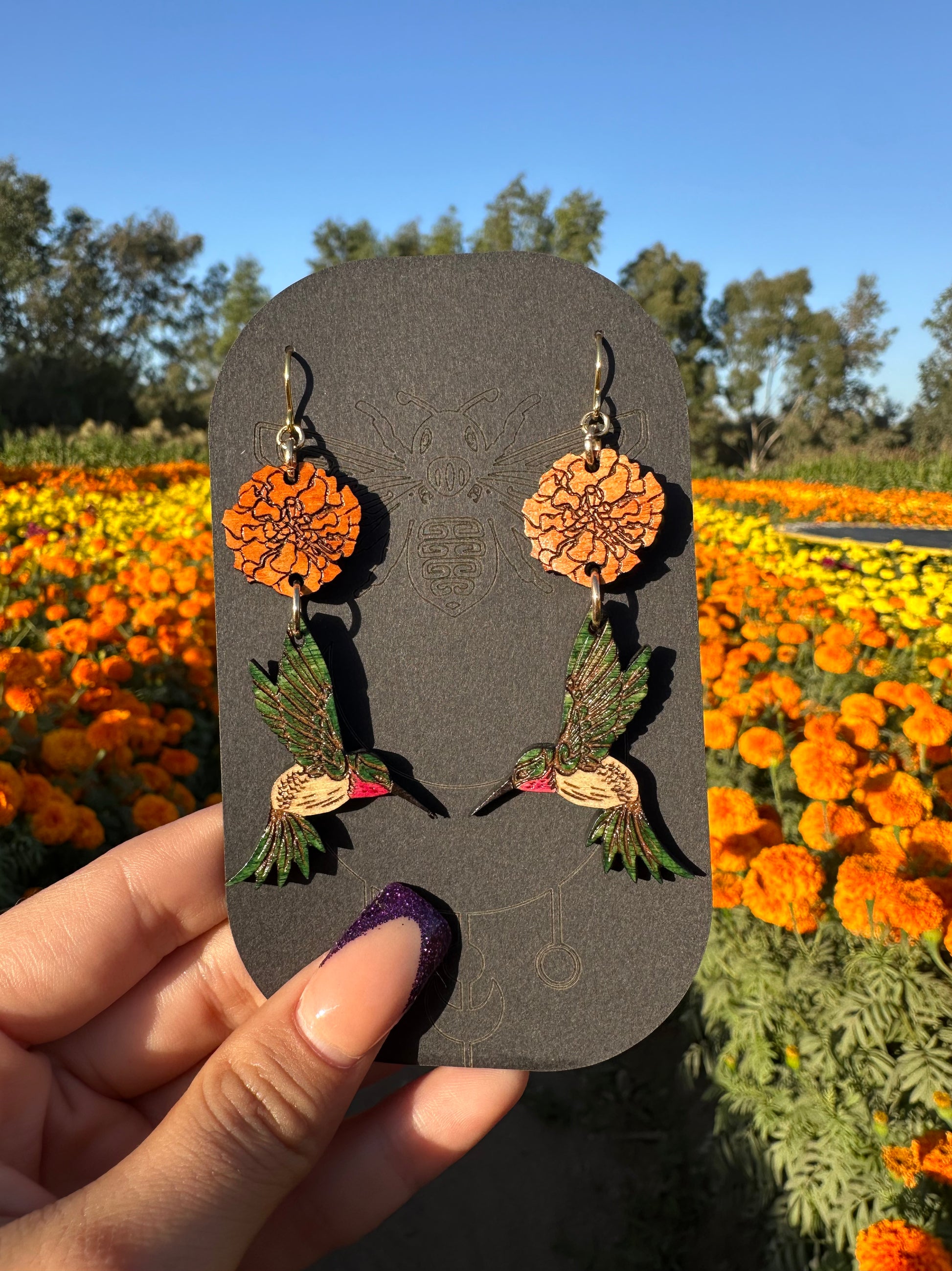 wood engraved earrings of an orange marigold with a green/white hummingbird with pink under beak. Earrings are on a gold colored hypoallergenic hook. Earrings are being held on a black cardstock earring holder with a background of orange and yellow marigold fields. 