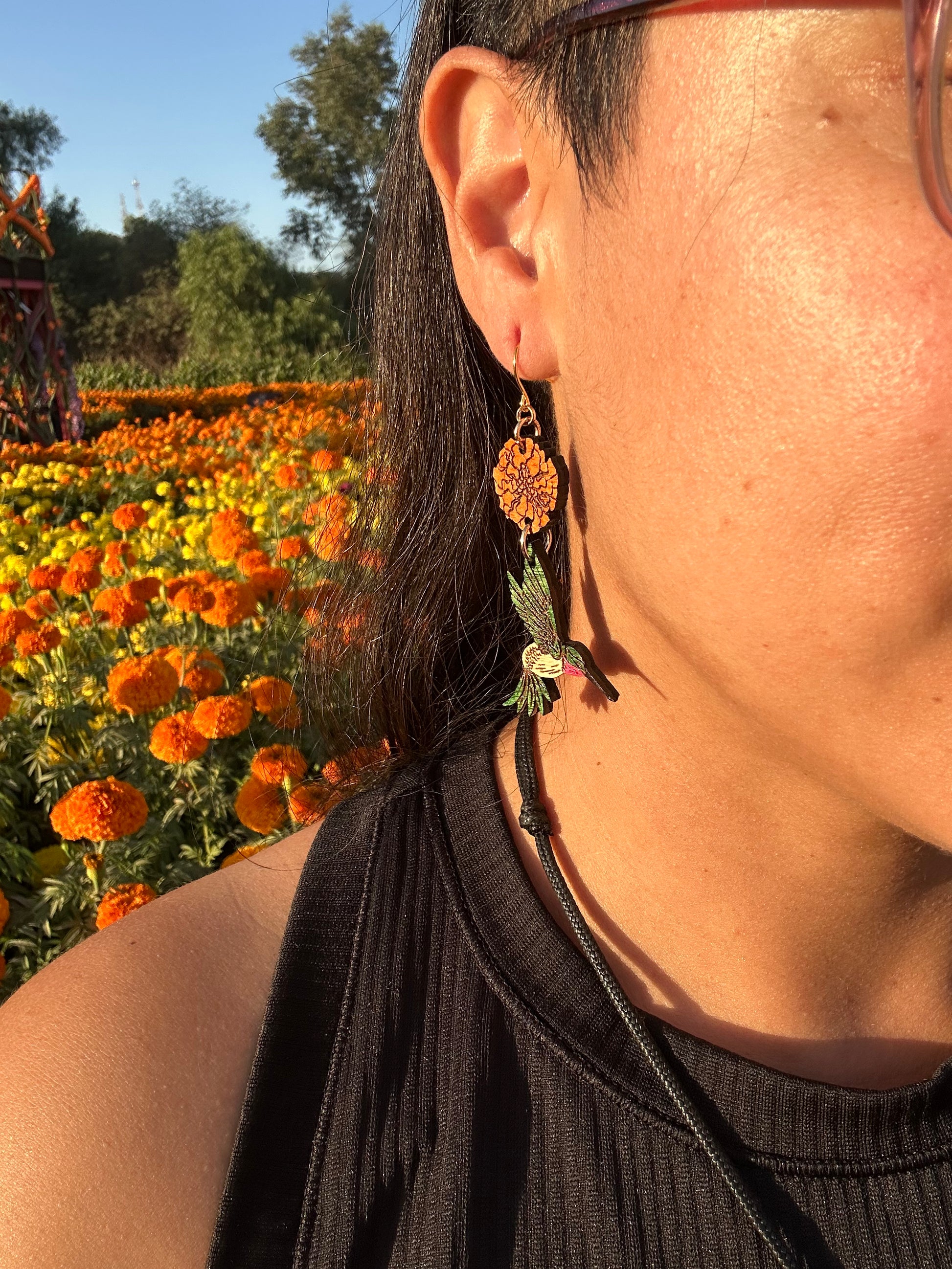 wood engraved earrings of an orange marigold with a green/white hummingbird with pink under beak. Earrings are on a gold colored hypoallergenic hook. Earrings are being worn by a women with closeup of side profile and zoomed in on the ear with a background of orange and yellow marigold fields. 