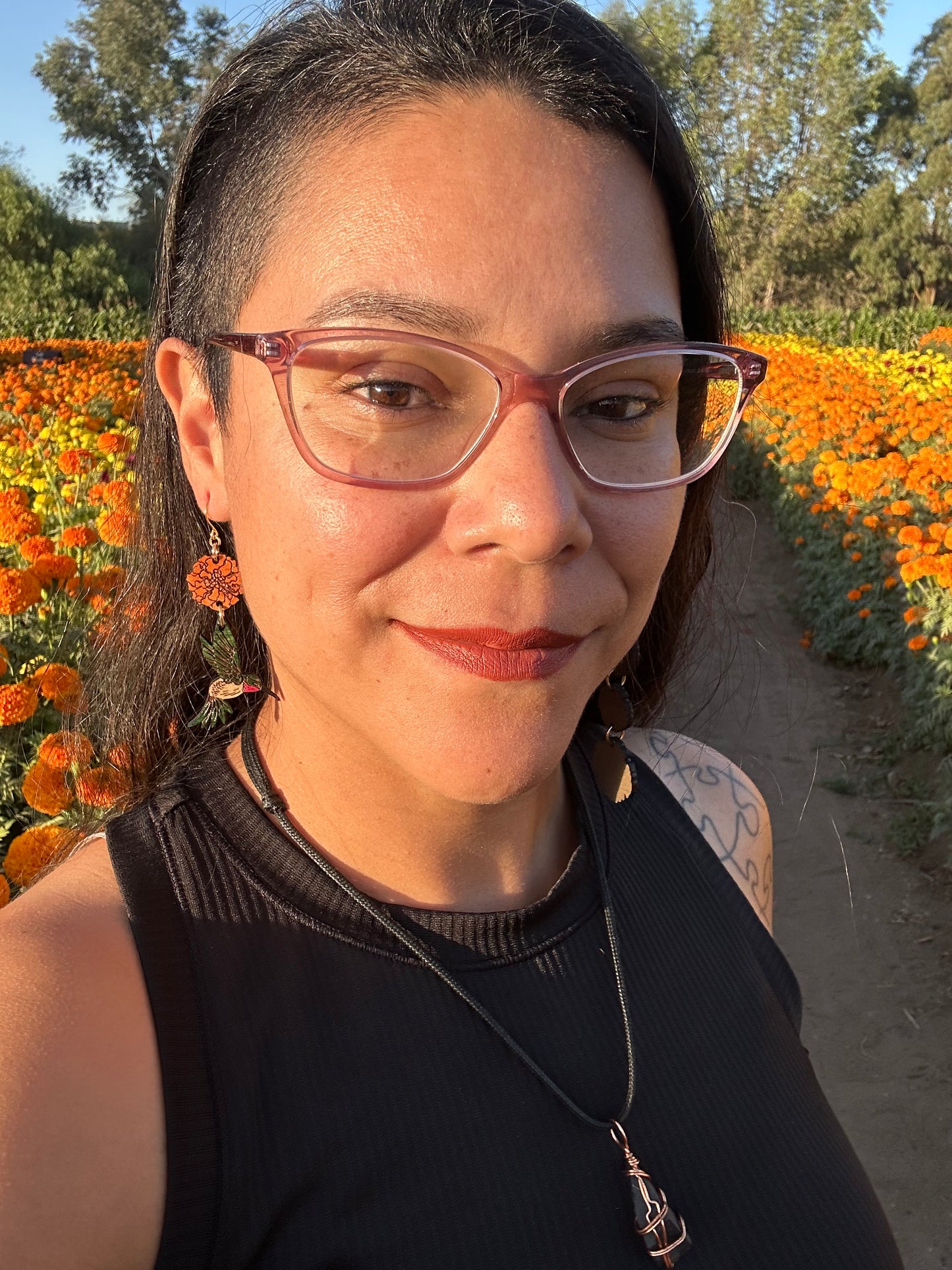 wood engraved earrings of an orange marigold with a green/white hummingbird with pink under beak. Earrings are on a gold colored hypoallergenic hook. Earrings are being worn by a women wearing glasses with a background of orange and yellow marigold fields. 