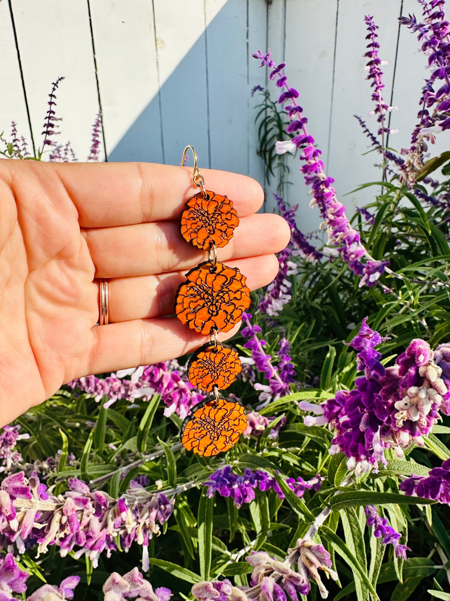 wood engraved orange marigold 4 strand of different sizes earrings on gold colored hypoallergenic hooks. earrings placed on hand to showcase the length with background of a purple mexican sage bush. 