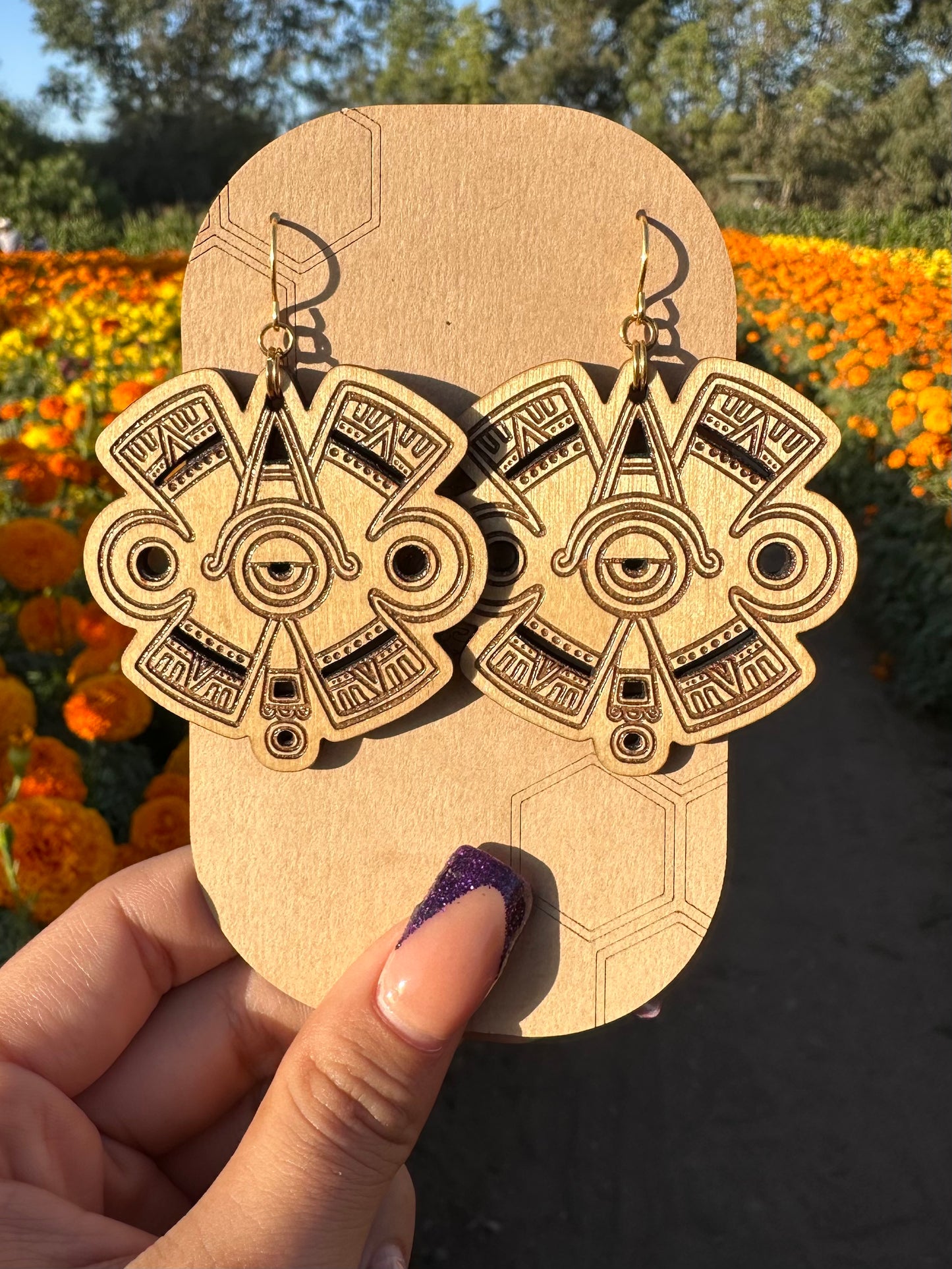 Ollin Earrings wood toned on gold colored hypoallergenic hooks. Earrings on brown card stock earring holder. Background of earrings is an orange and yellow marigold field. 