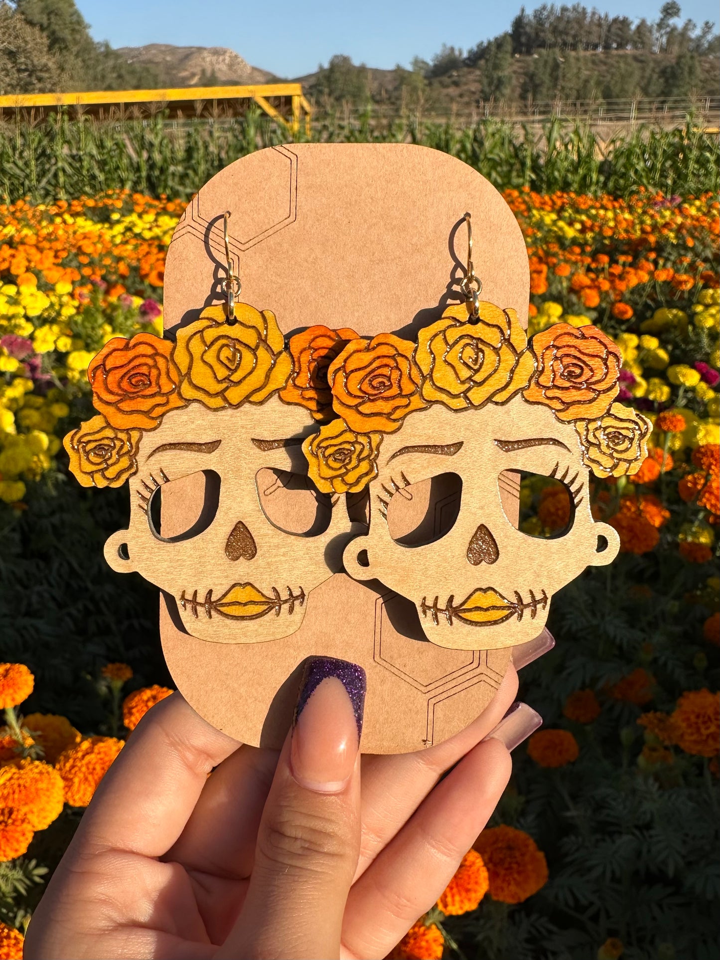 wood engraved catrina earrings with orange and yellow roses on head with gold colored hypoallergenic earring hooks on a brown card stock earring holder with a background of a marigold field. 