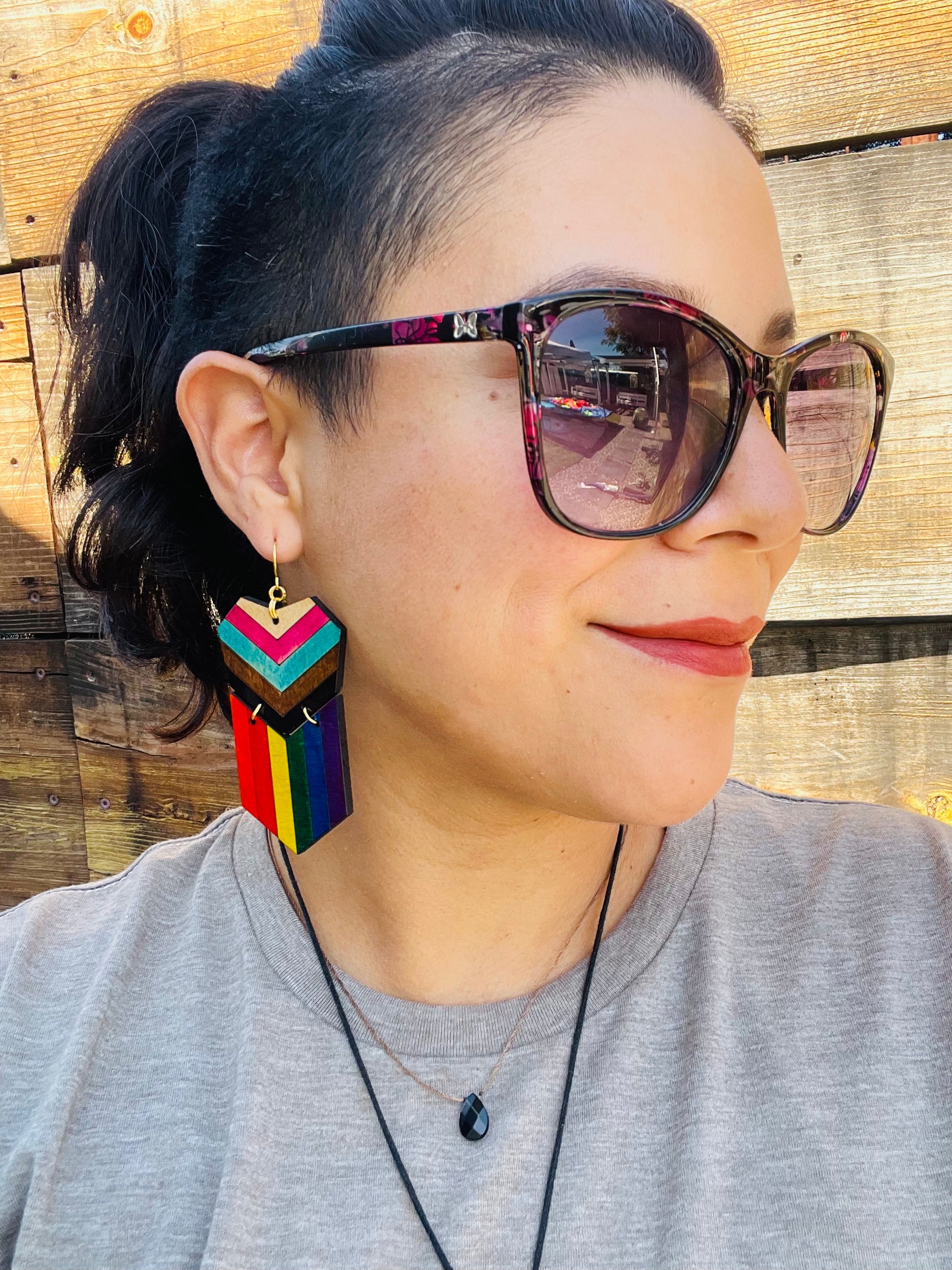 wood engraved progress pride earrings with a gold colored hypoallergenic hook. Worn by a woman with hair in ponytails and big sunglasses. 