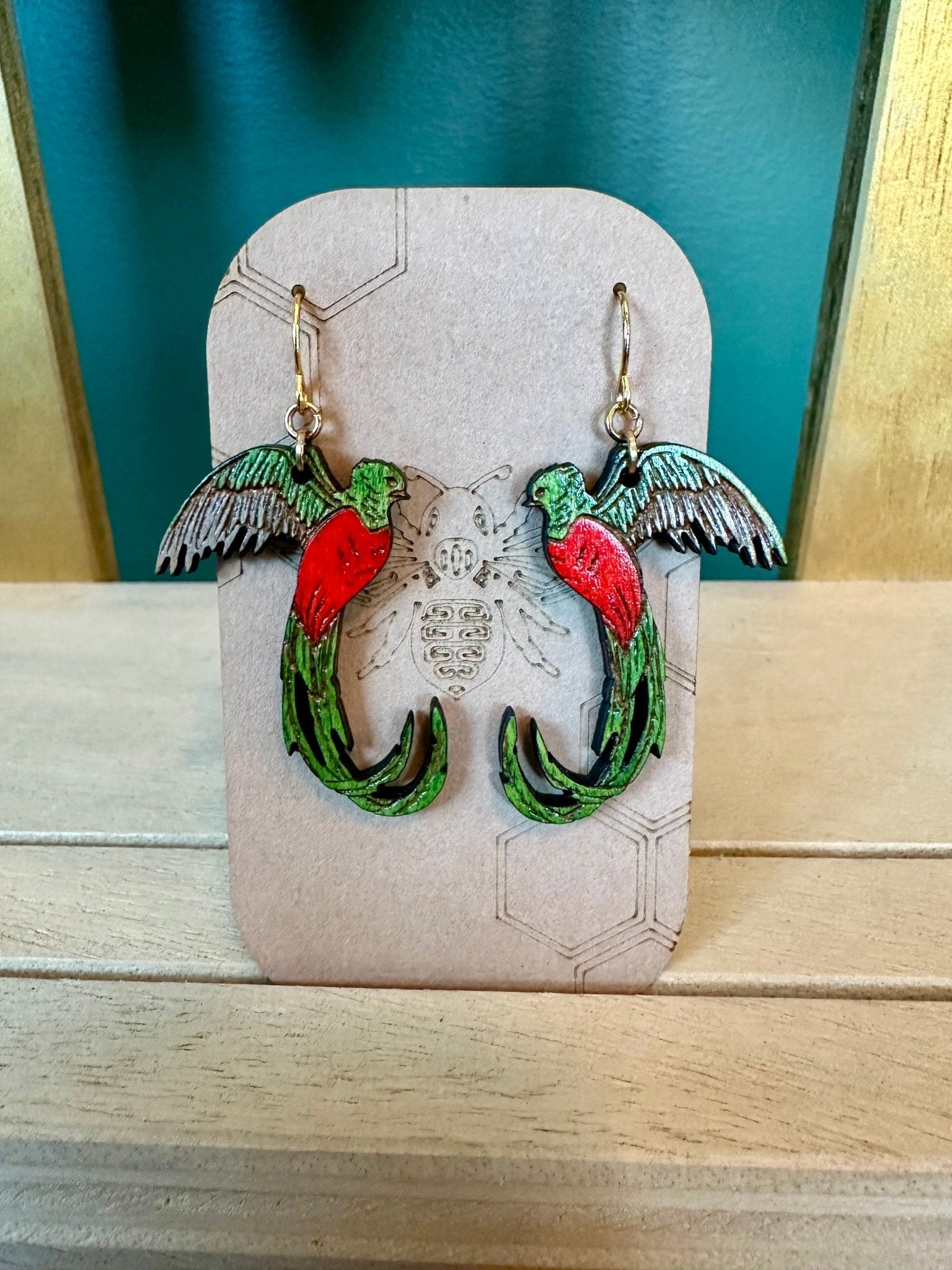 quetzal earrings wood engraved green and red bird on a gold colored hypoallergenic hook. earrings on a brown card stock earring holder. 