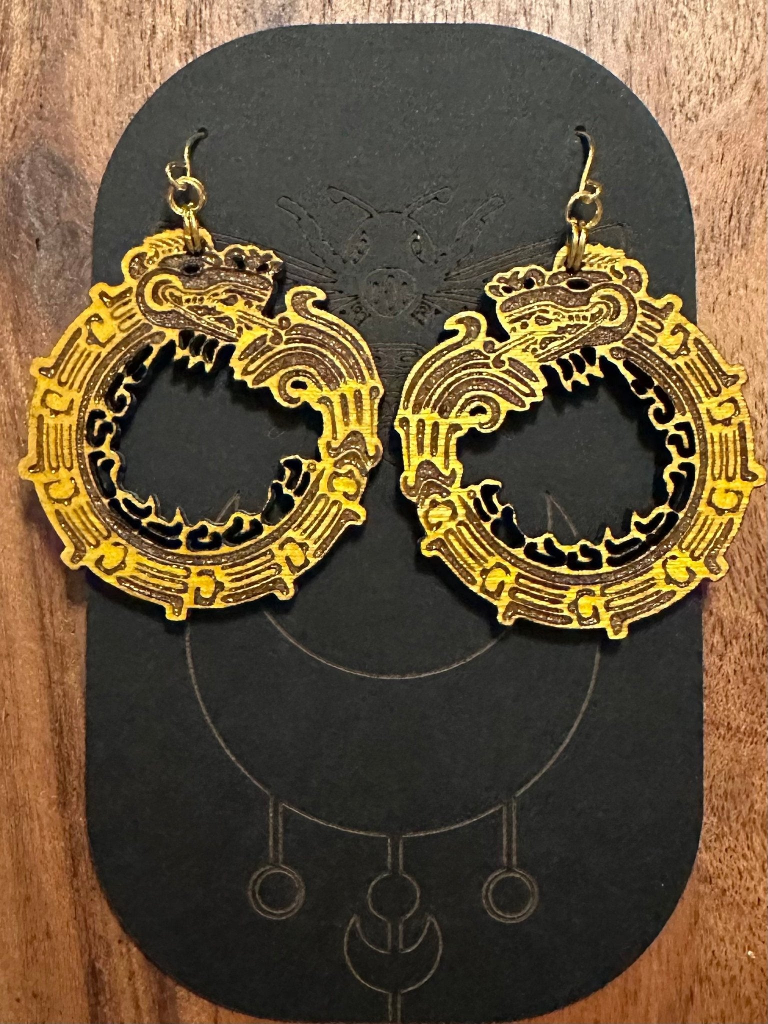 wood engraved yellow Quetzalcoatl Earrings on gold colored hypoallergenic hooks. earrings on a black card stock earring holder.