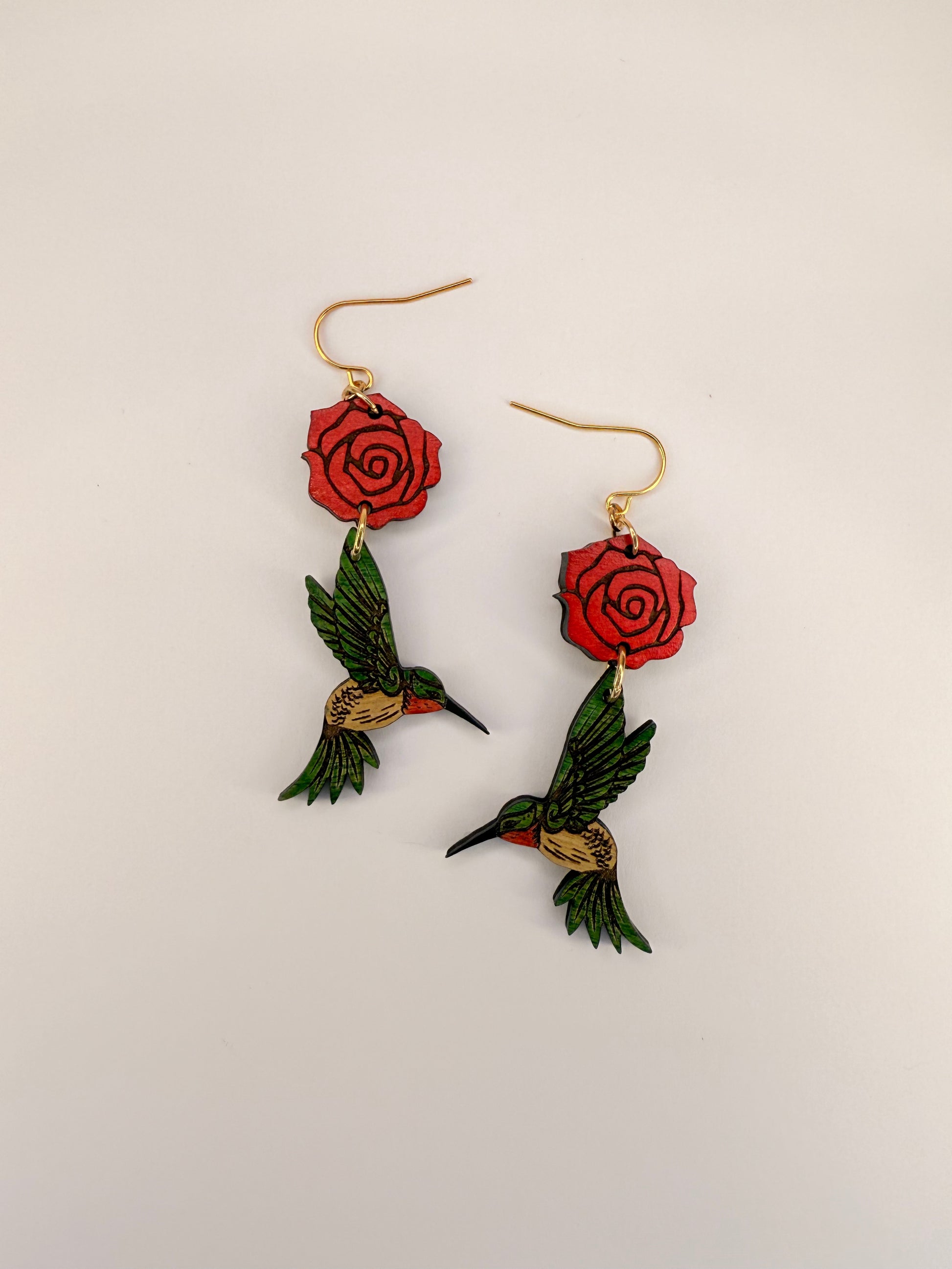 Pair of earrings with red roses and green hummingbirds on a beige background.