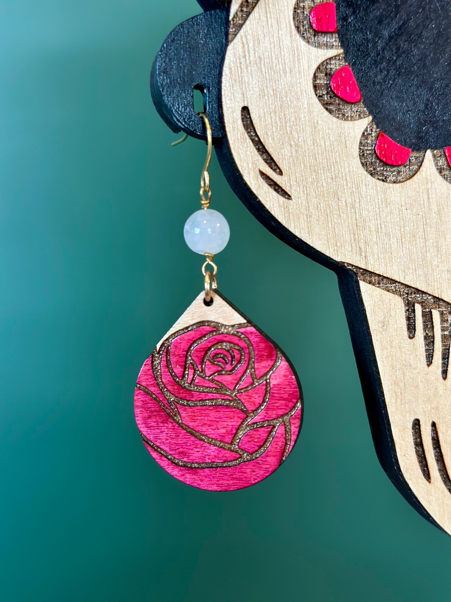 wood engraved pink colored rose teardrop shaped earrings with a rose quartz crystal bead on gold colored hypoallergenic earring hooks worn by a wood engraved catrina skeleton woman display close up.