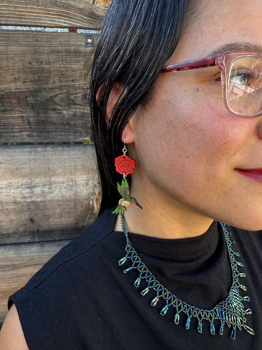 Person wearing red rose and green hummingbird earrings and a black top with decorative elements on a wooden surface.