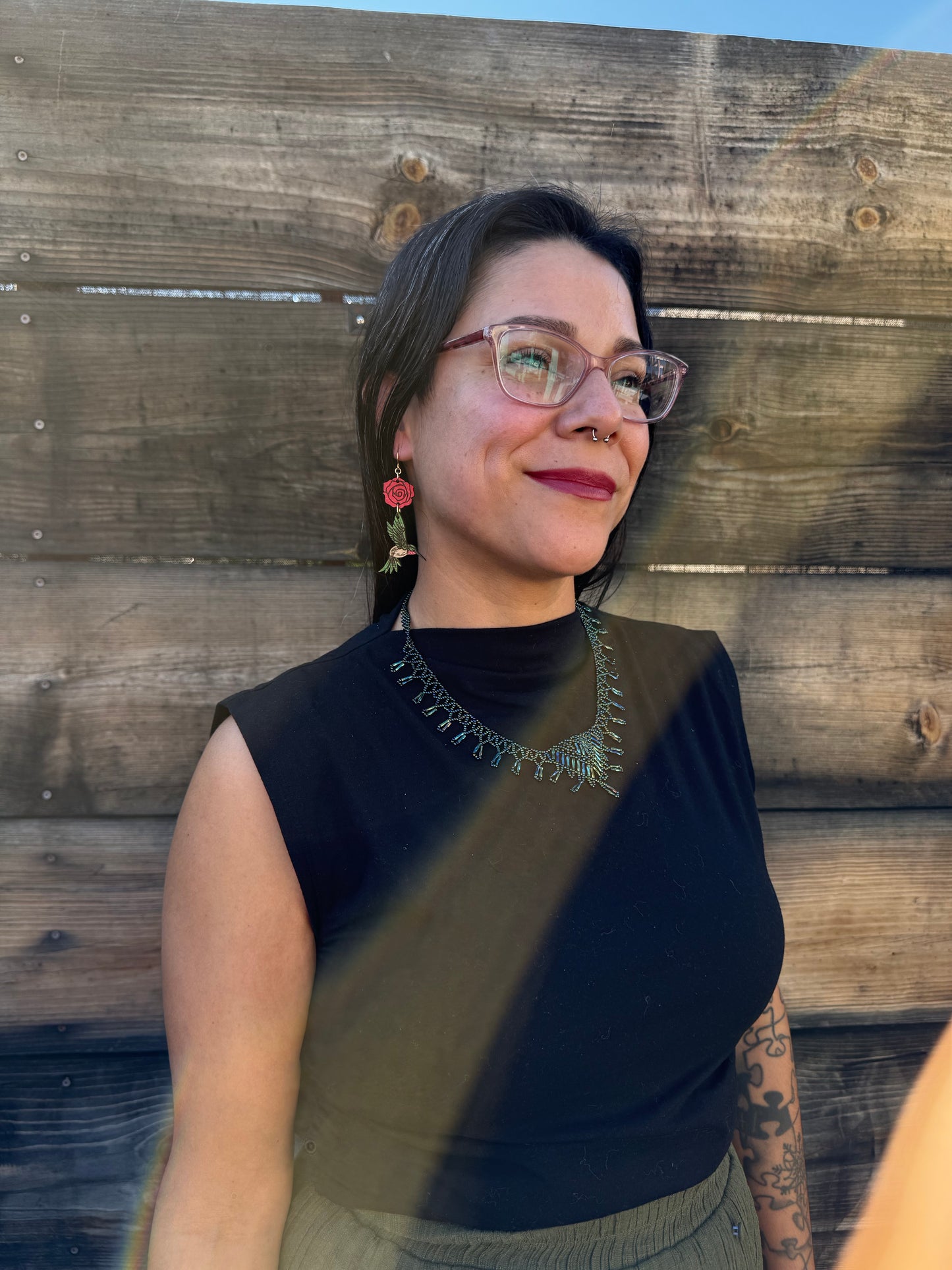 Person wearing red rose and green hummingbird earrings and  a black shirt with a decorative neckline against a wooden background.