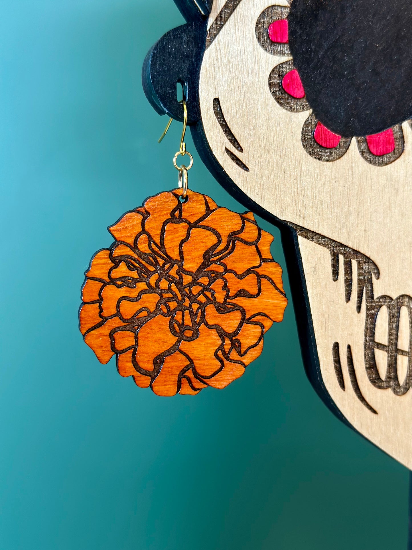 large wood engraved orange marigold earring on gold colored hypoallergenic earring hook placed on earring hole of a wood engraved catrina model close up. 