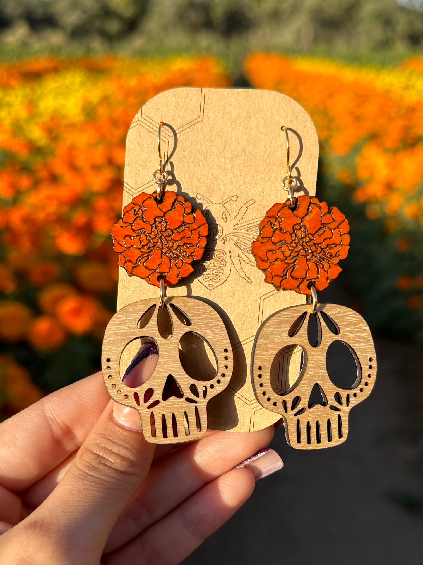 wood engraved orange marigold with sugar skull cutout earrings with gold colored hypoallergenic earring hooks on a brown card stock earring holder with a background of a marigold field. 