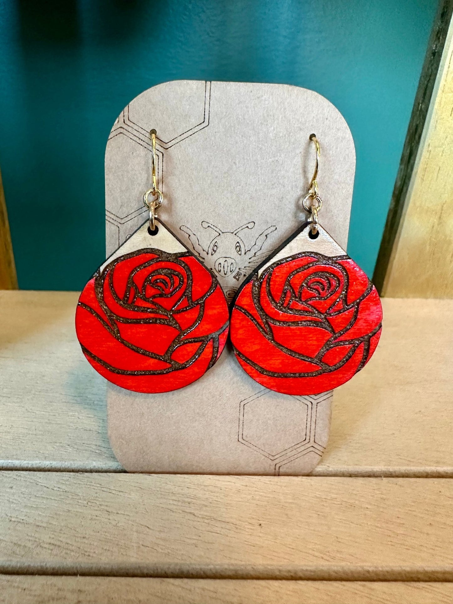 wood engraved red colored rose teardrop shaped earrings on gold colored hypoallergenic earring hooks. earrings on a brown card stock earring holder. 