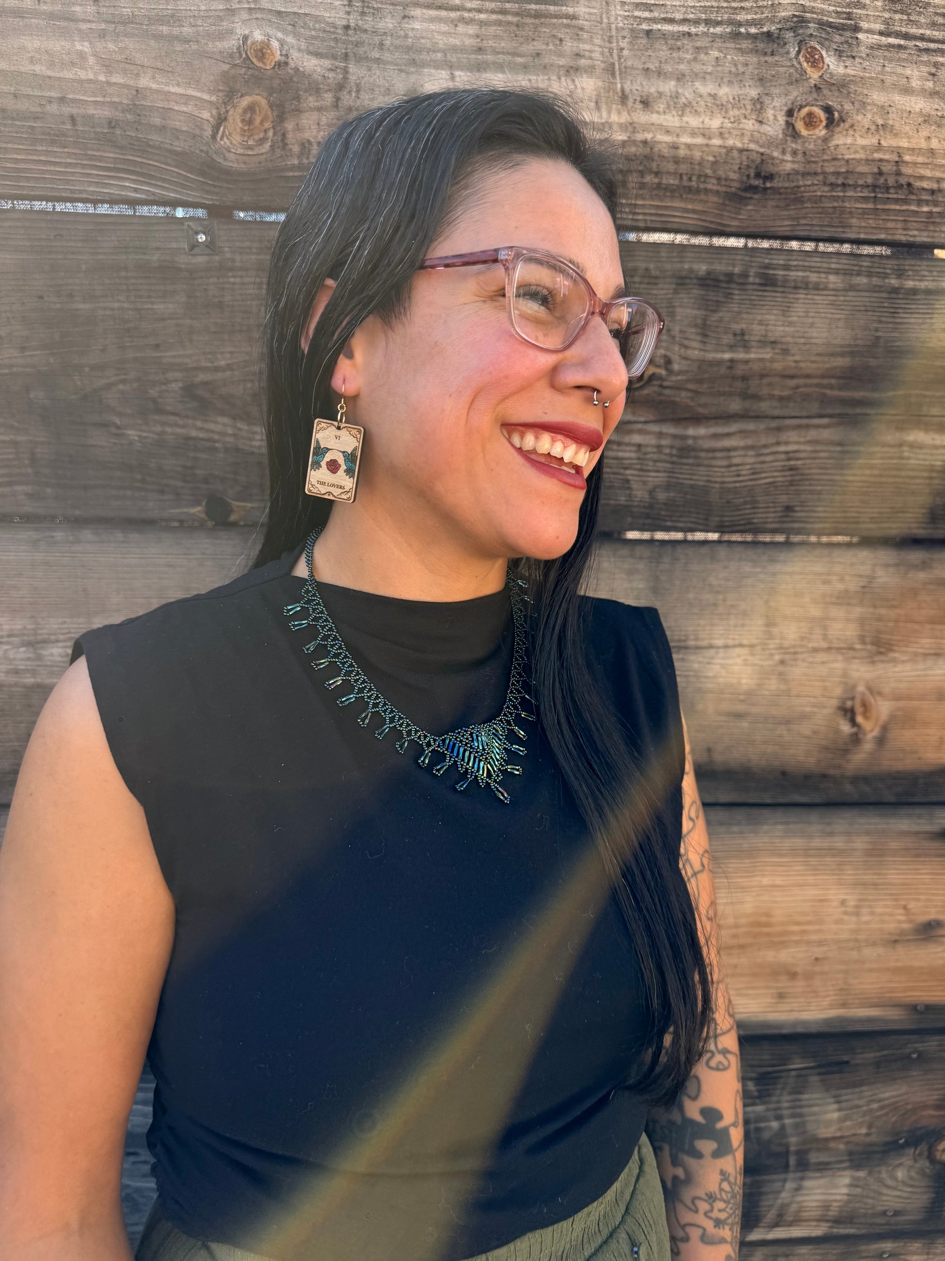 A woman smiling wearing the lovers tarot with an engraving of hummingbirds and roses.