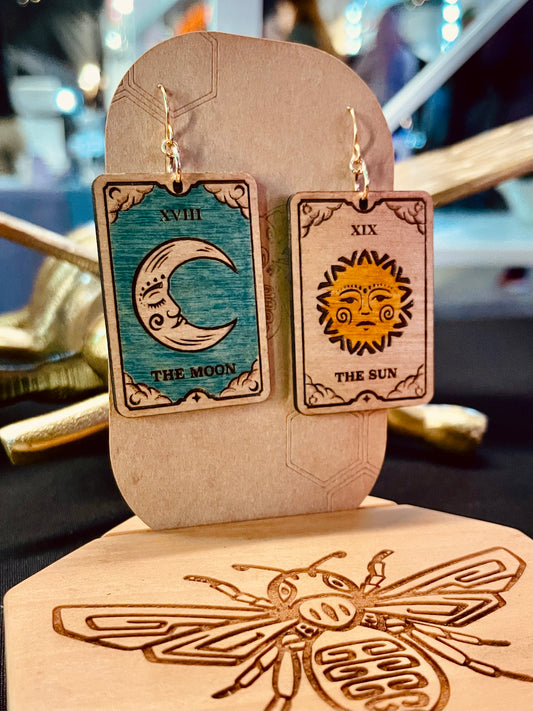 wood engraved tarot card earrings. One shows a crescent moon with a blue background and the other shows a yellow colored sun. These earrings show two different designs combined as a pair of earrings. These earrings have a gold colored hypoallergenic nickel free hook and are displayed on a brown cardstock earring holder. 