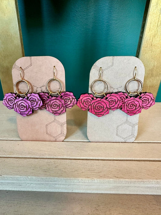 wood engraved purple colored rose earrings on a gold colored circle hoop on a gold colored hypoallergenic earring hook. wood engraved pink colored rose earrings on a gold colored circle hoop on a gold colored hypoallergenic earring hook. Pairs of earrings on brown card stock earring holders. 