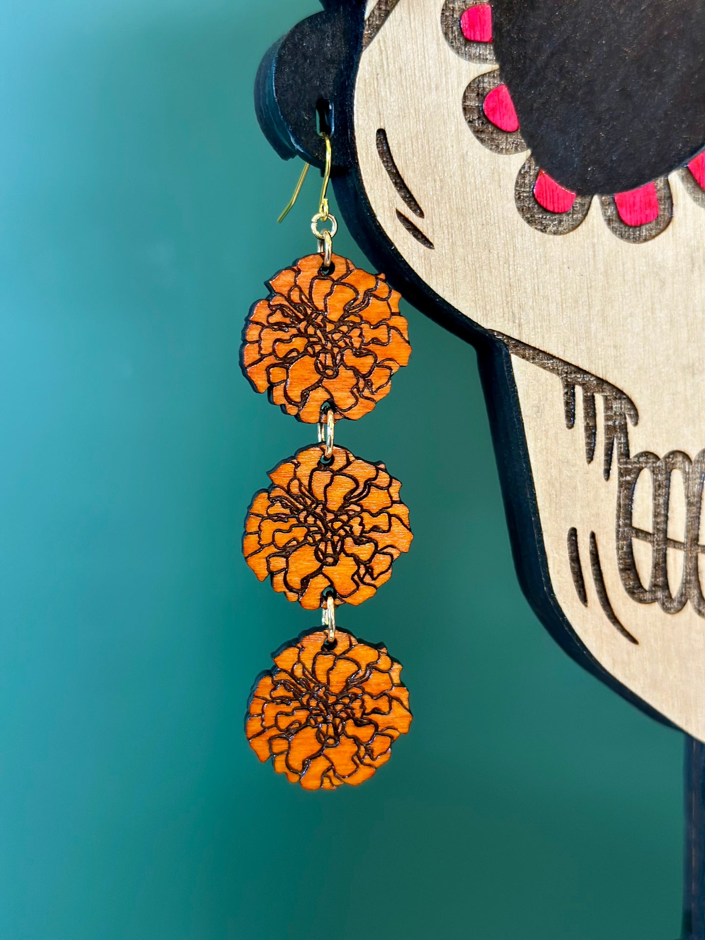 three layered wood engraved orange marigold earring on gold colored hypoallergenic earring hook placed on earring hole of a wood engraved catrina model close up. 